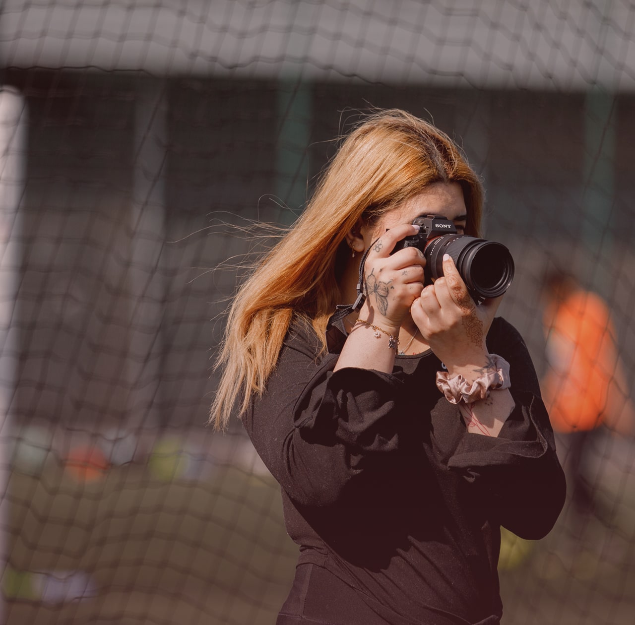 The image shows a person with long hair holding a camera, taking a photo. They're wearing a black outfit and have some visible tattoos on their hand. The background is blurred, and there's a net visible, indicating they might be near a sports field.