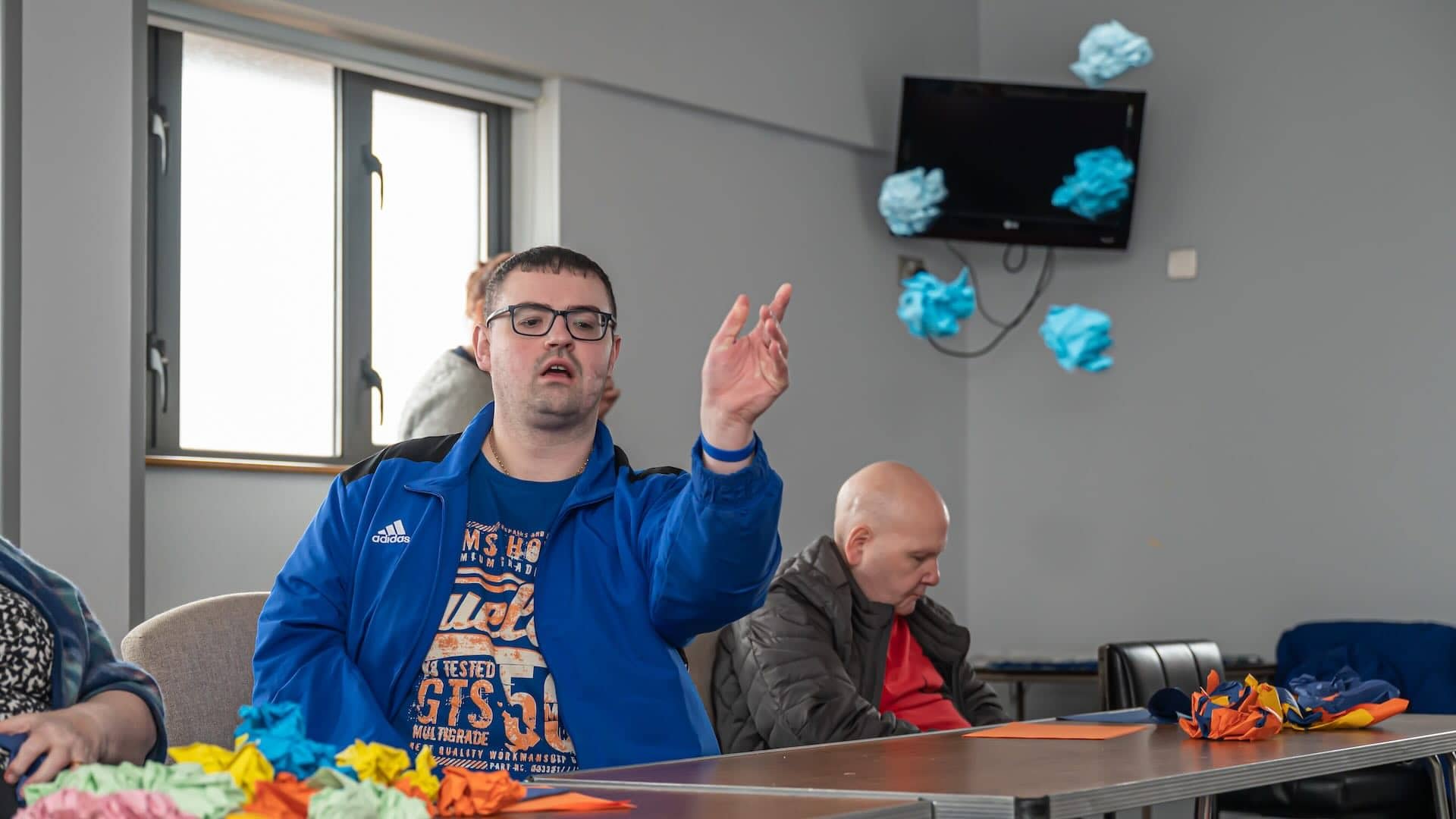 The image shows a man wearing glasses and a blue jacket in an indoor setting with other people. He appears to be engaging in an activity with colorful paper or fabric on the table. There are crumpled blue pieces on a table and some also floating