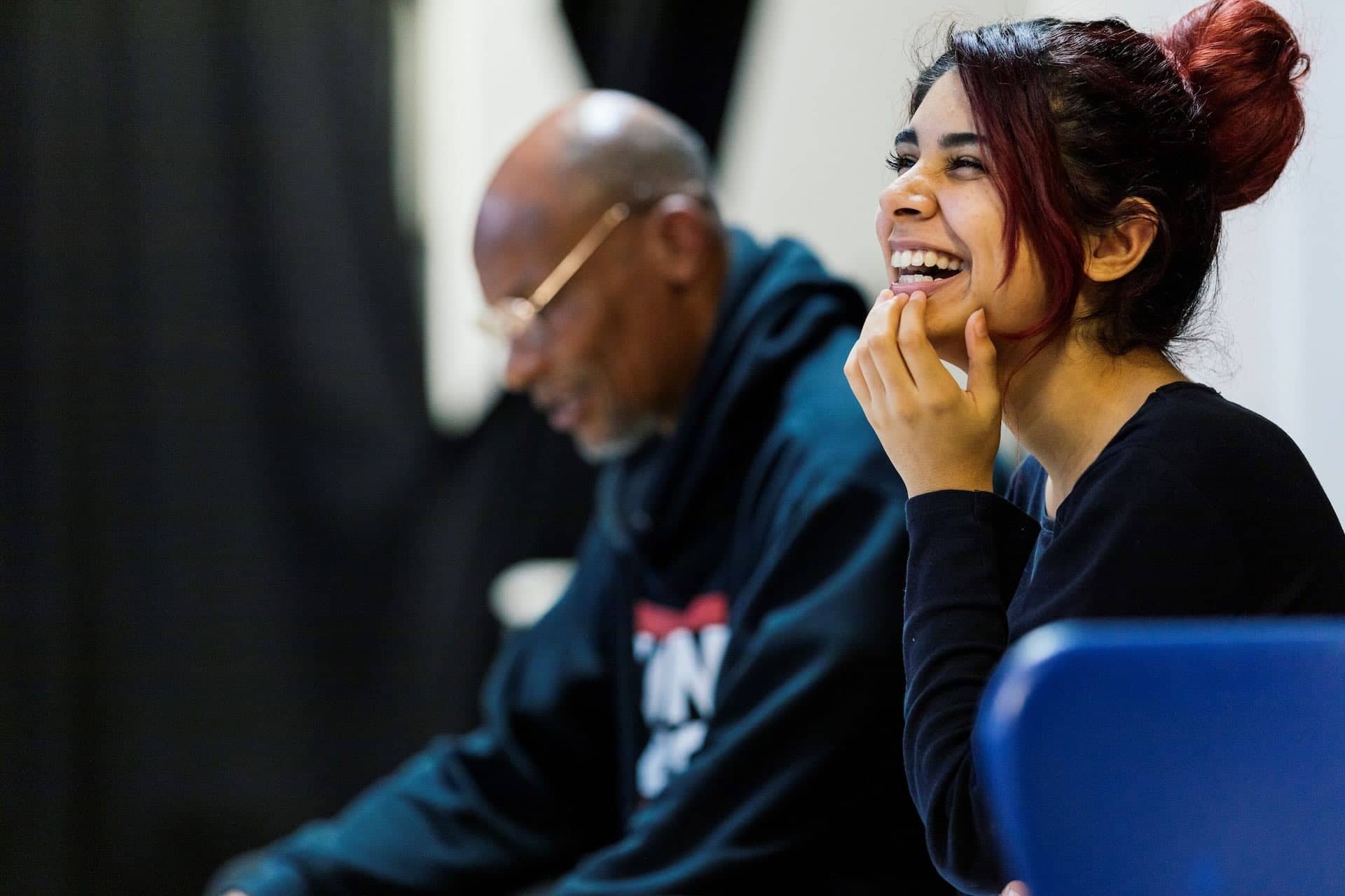 The image shows two people. A woman is smiling and appears to be engaged or in conversation. In the background, a man wearing glasses is also present. The scene seems casual and pleasant, with the focus on the woman's expression.