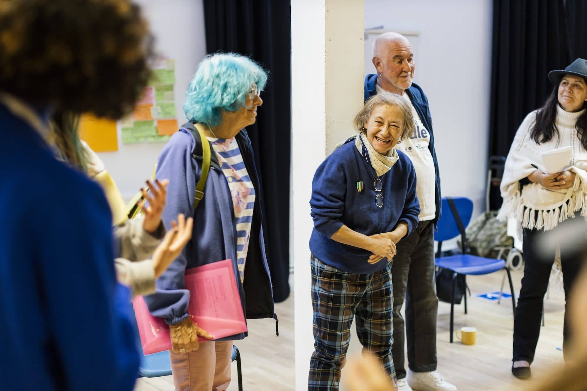 The image shows a group of people standing in a room, seemingly engaged in a conversation or activity. There are several chairs around, and a few people are holding papers or notepads. The atmosphere appears casual and friendly. One person is leaning on
