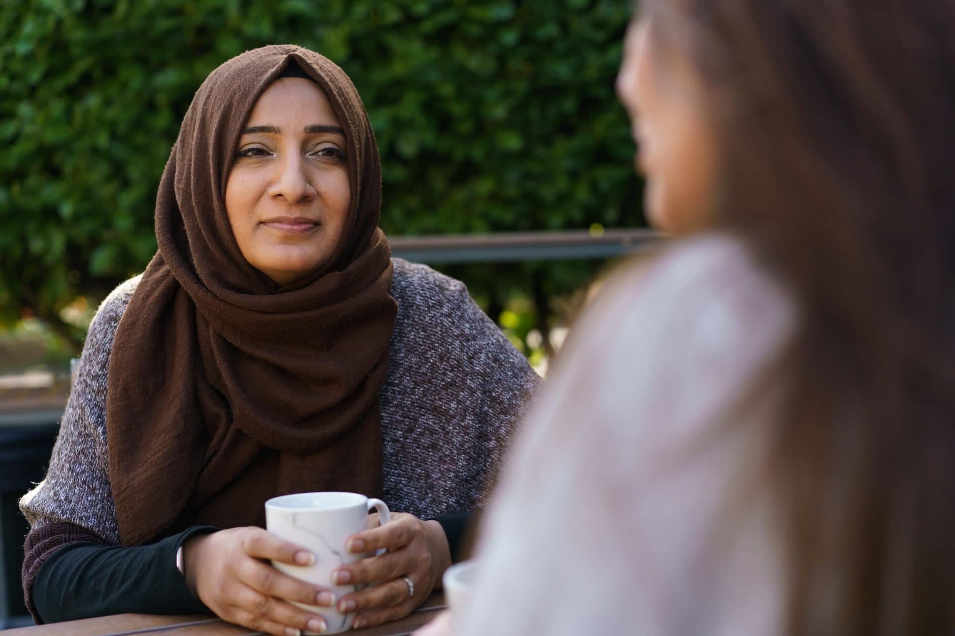 The image shows two people sitting outdoors, likely at a café or similar setting. One person is wearing a brown hijab and holding a white mug, looking across at the other person, who is partially visible and out of focus. There are green shrubs