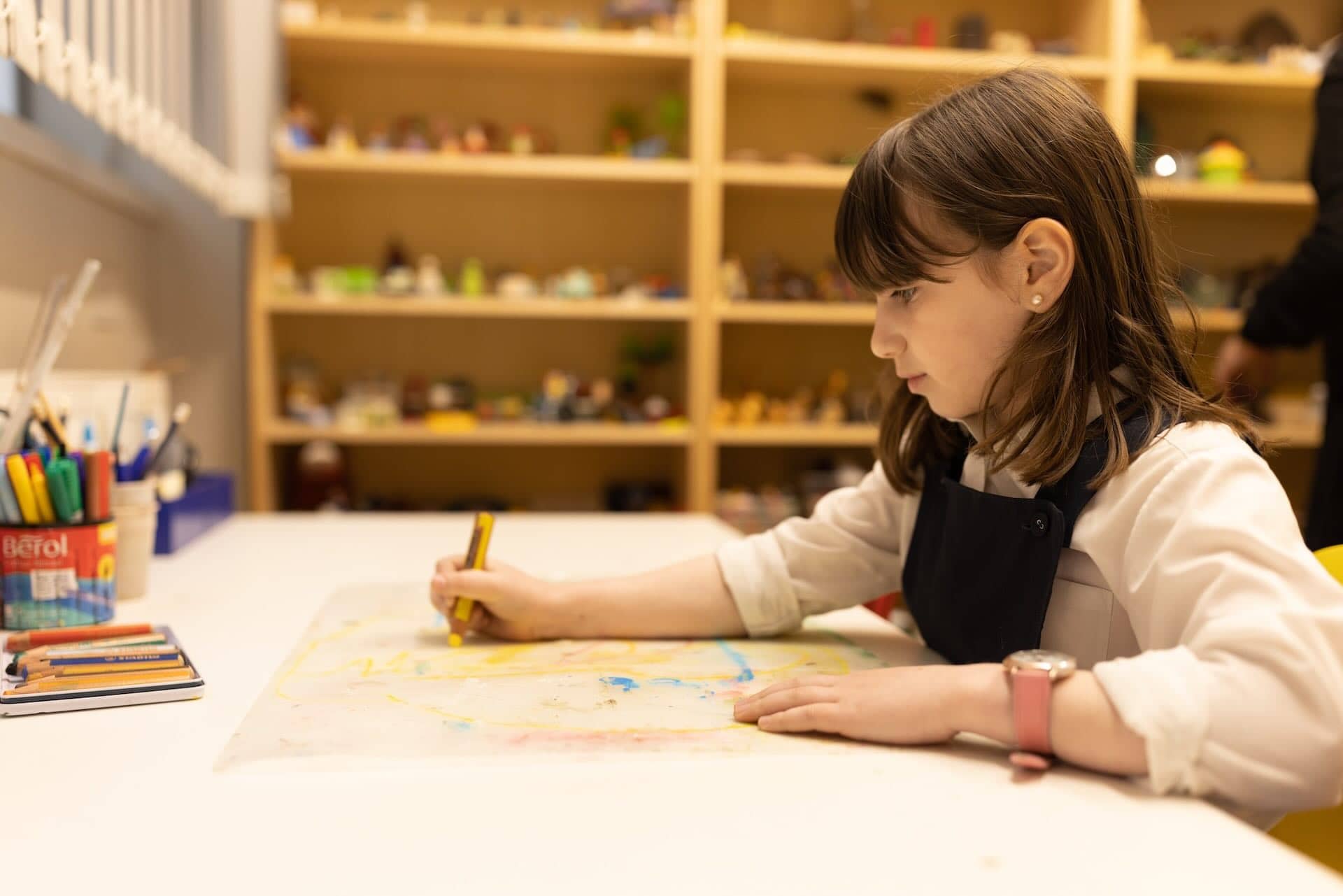 The image shows a young child engaged in drawing or coloring at a table. The child is holding a colored pencil or crayon, working on a piece of paper. There are art supplies, like colored pencils and markers, on the table. In the