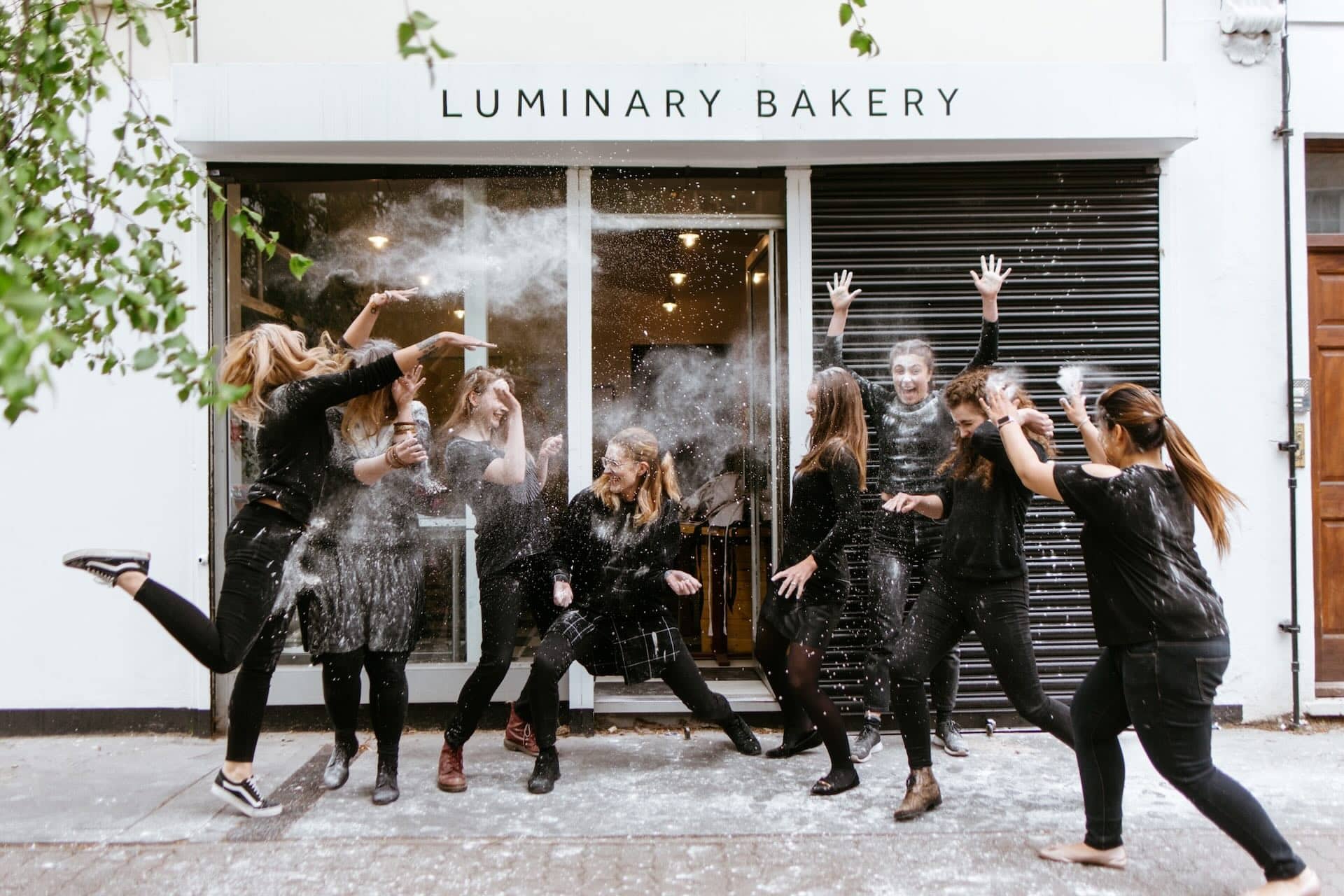 The image shows a group of people outside a place called LUMINARY BAKERY. They appear to be having fun, throwing flour at each other. The group is wearing casual clothing, and there is flour in the air and on the ground
