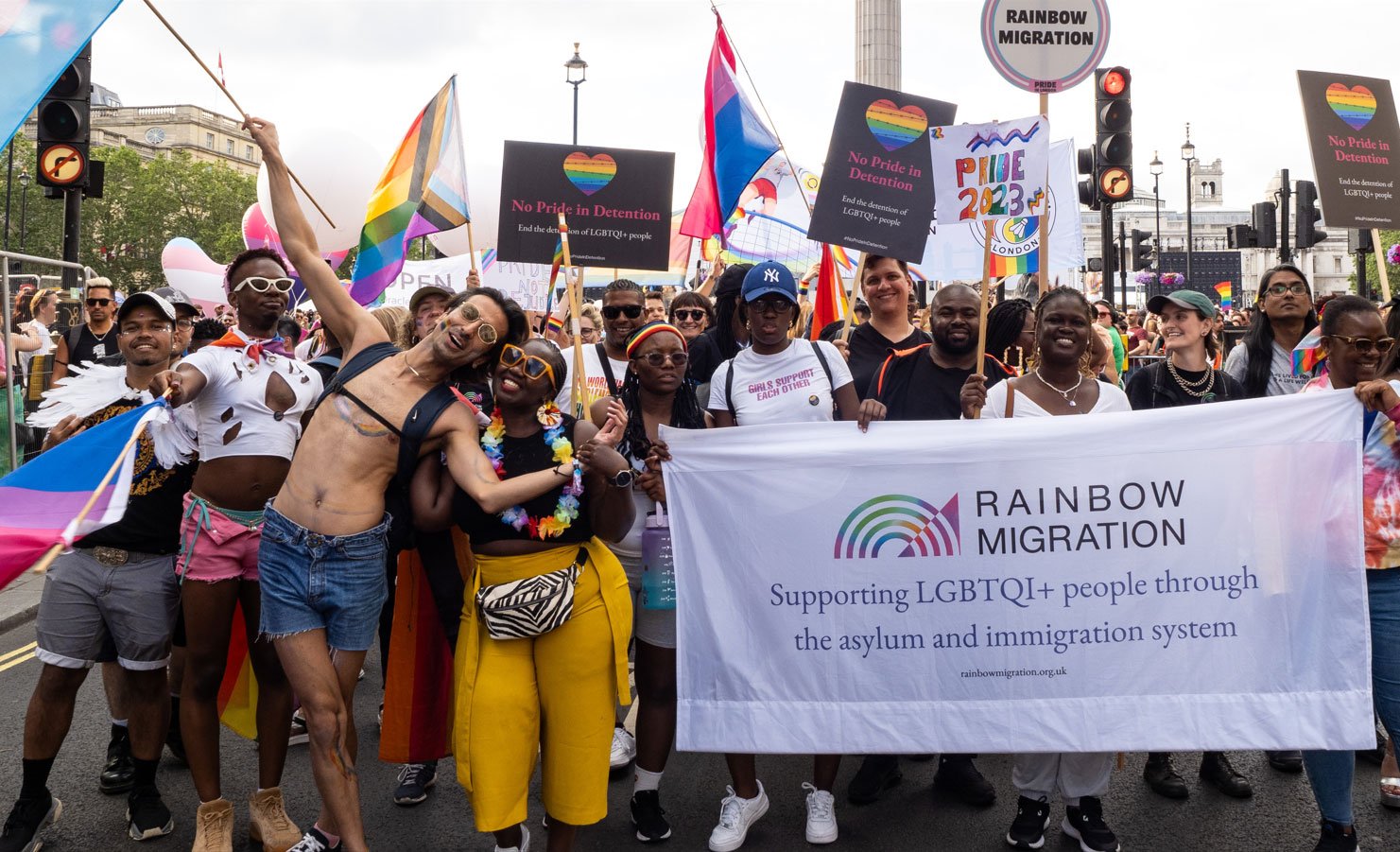 A group of young migrant people march in a pride parade in London, wearing rainbow face paint and holding Pride signs and a large Rainbow MIgration banner