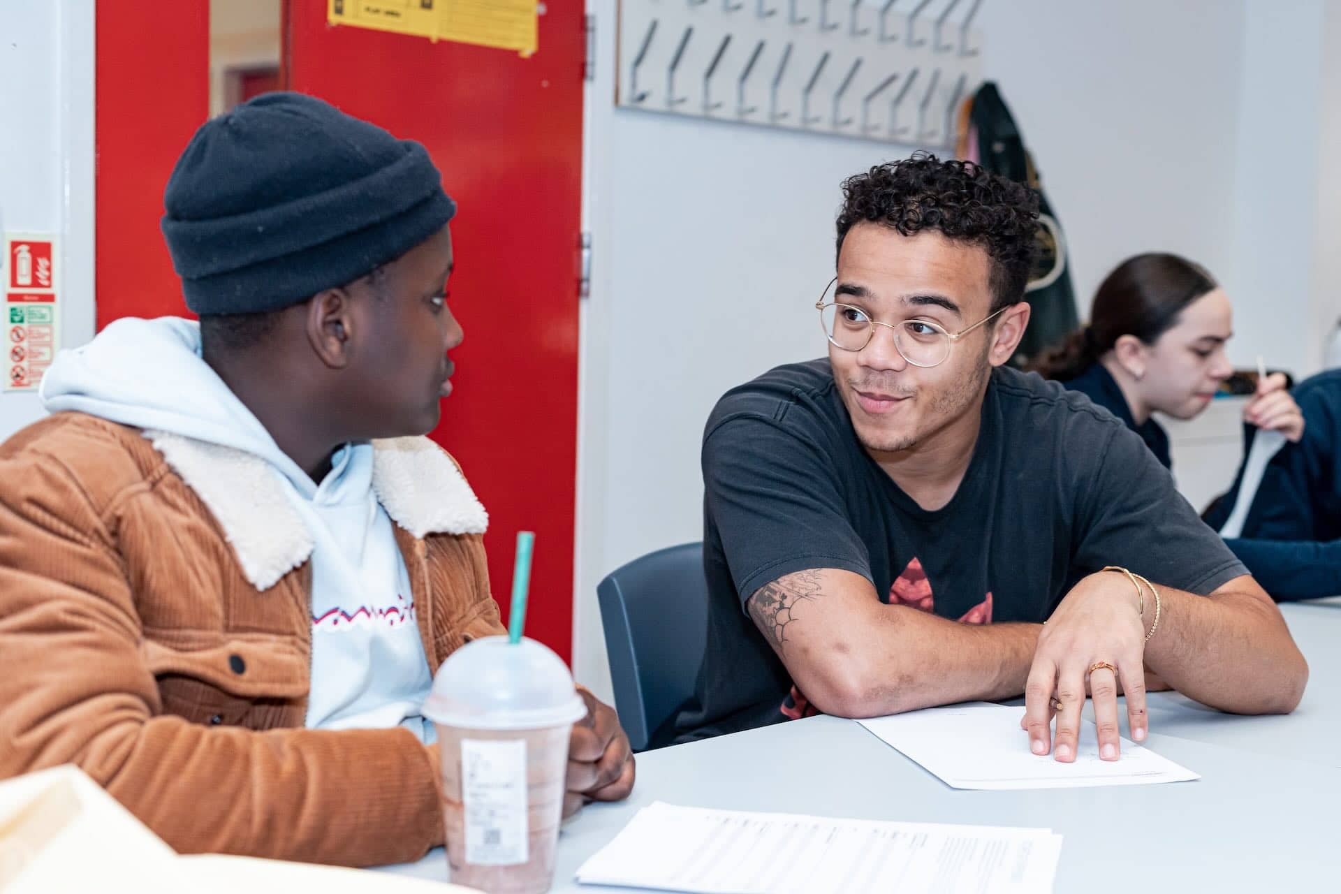 The image shows two people sitting at a table engaged in conversation. One person is wearing a brown jacket with a white hoodie and a black beanie, while the other is wearing a black t-shirt and glasses. There are papers on the table and a