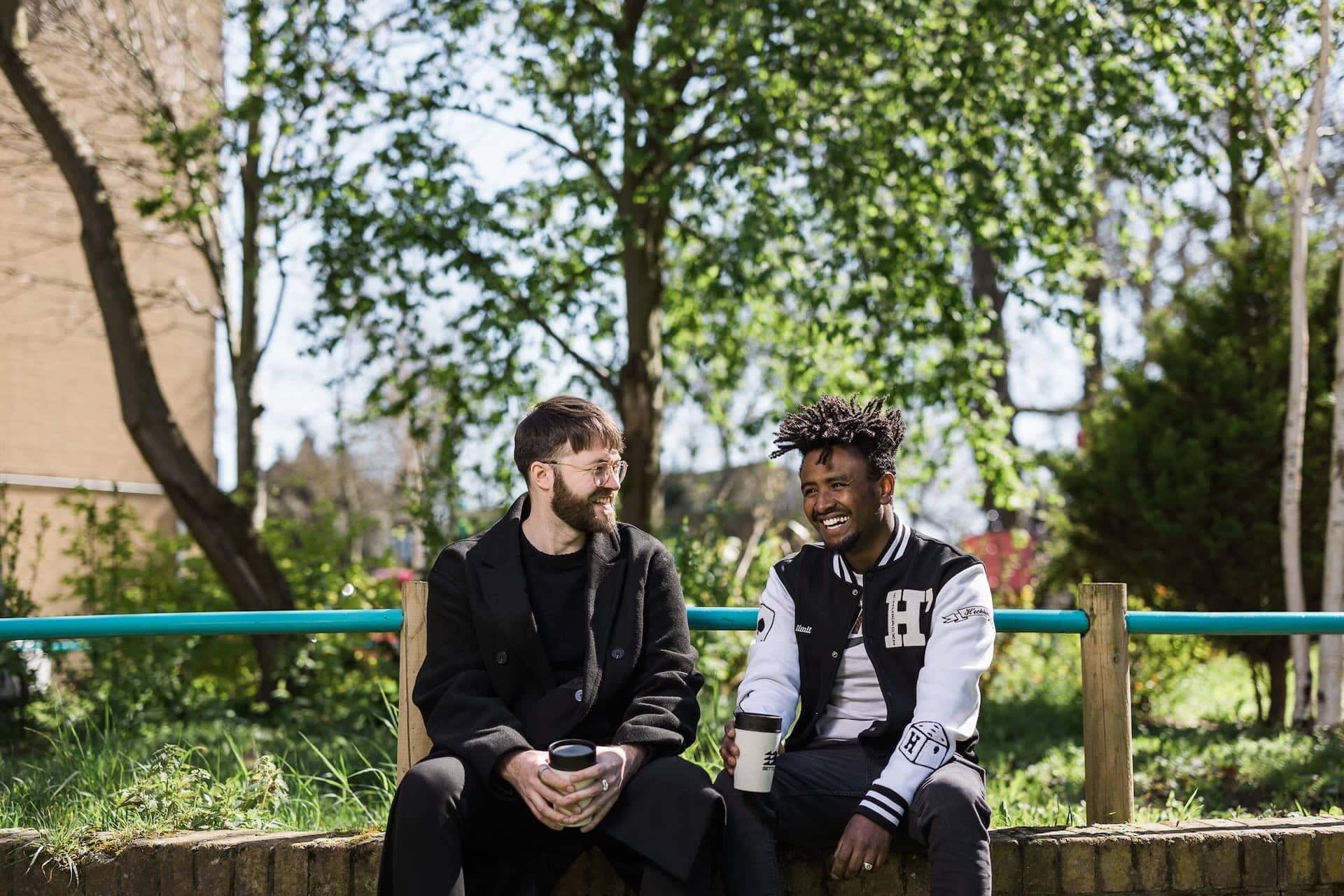 The image shows two people sitting outdoors on a low wall, having a conversation. They appear to be smiling and holding coffee cups. The setting includes green trees and grass in the background, suggesting it's a pleasant, sunny day.
