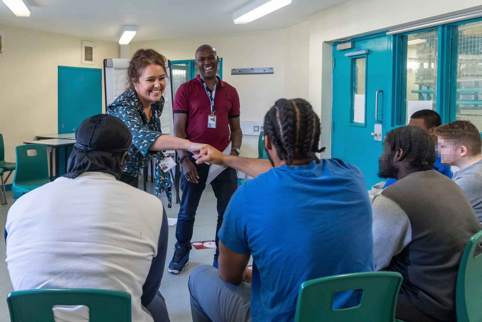 The image shows a group of people in a room. One person is cheerfully shaking hands with another, creating a friendly atmosphere. There are chairs and tables, with a flipchart in the background, suggesting a meeting or workshop. The setting appears to