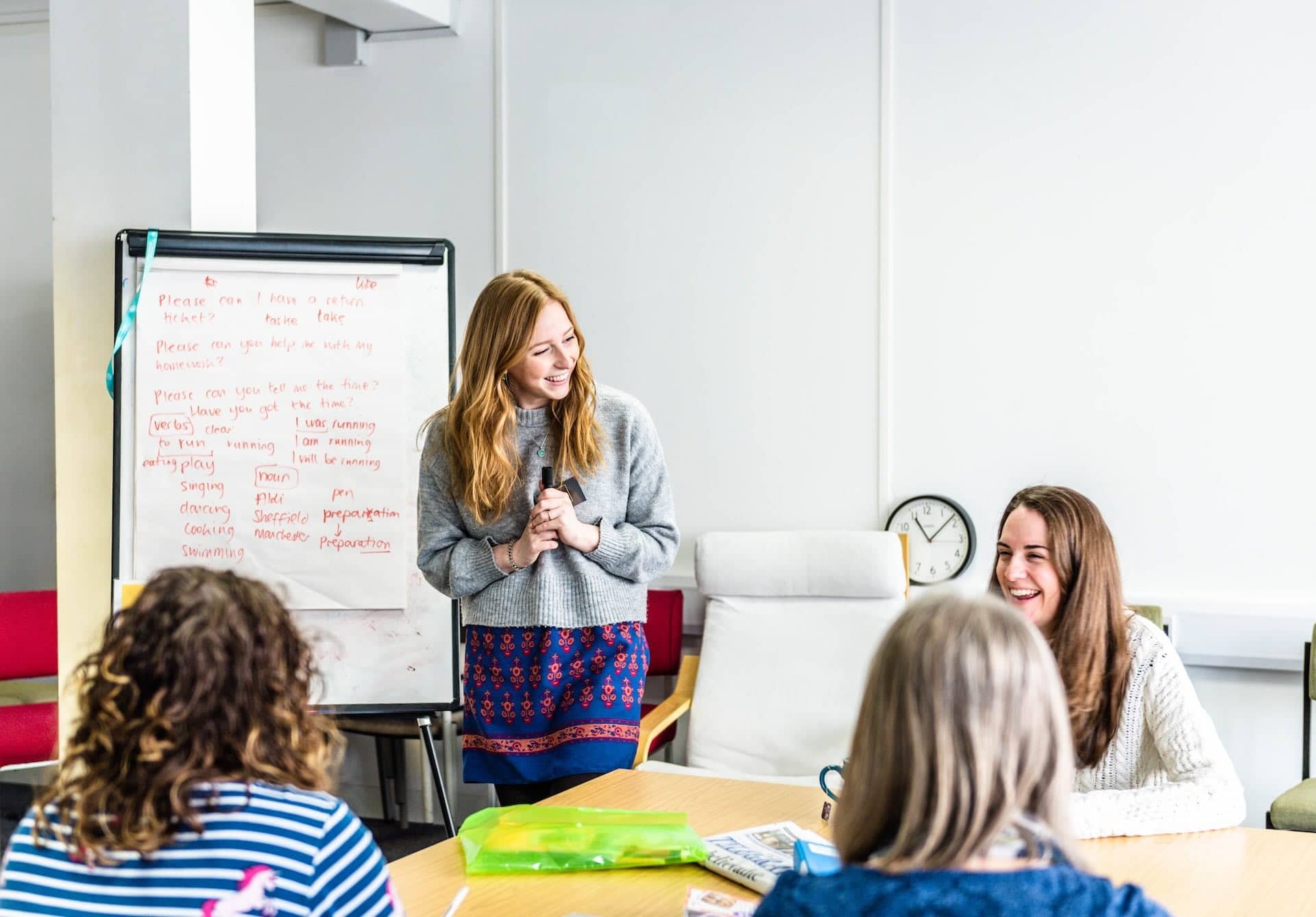 The image depicts a classroom or meeting setting with a group of people. A woman is standing near a flip chart with written notes and appears to be leading a discussion or giving a presentation. Several people are sitting around a table, engaged and smiling. The