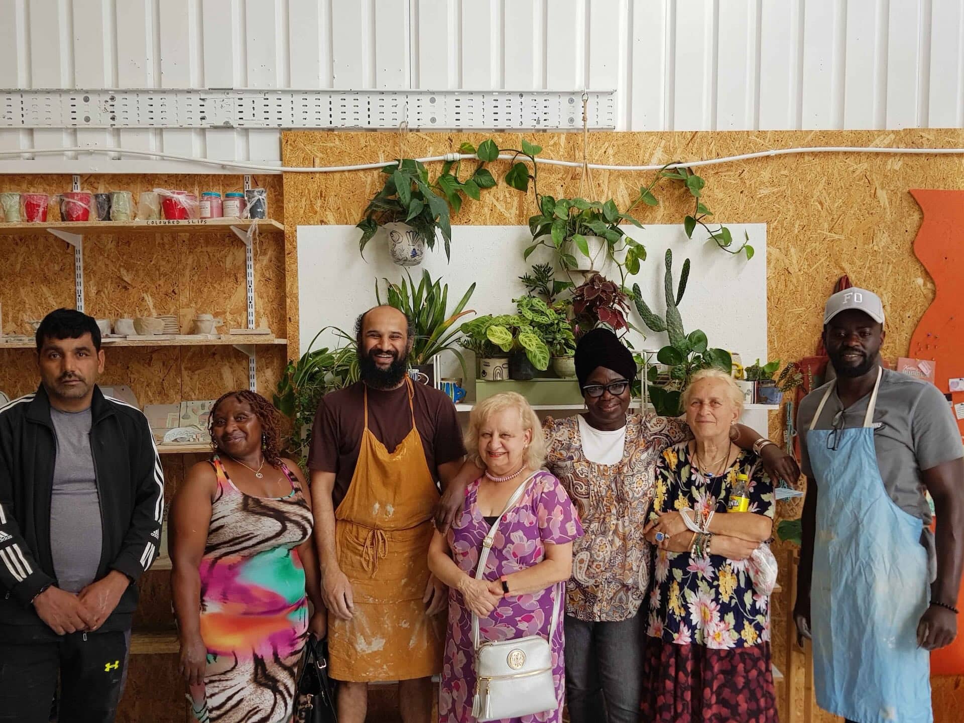 The image shows a group of seven people standing together in front of a background with shelves and plants. Some are wearing aprons, suggesting they might be in a workshop or studio setting, possibly related to crafts or pottery, given the presence of shelves with