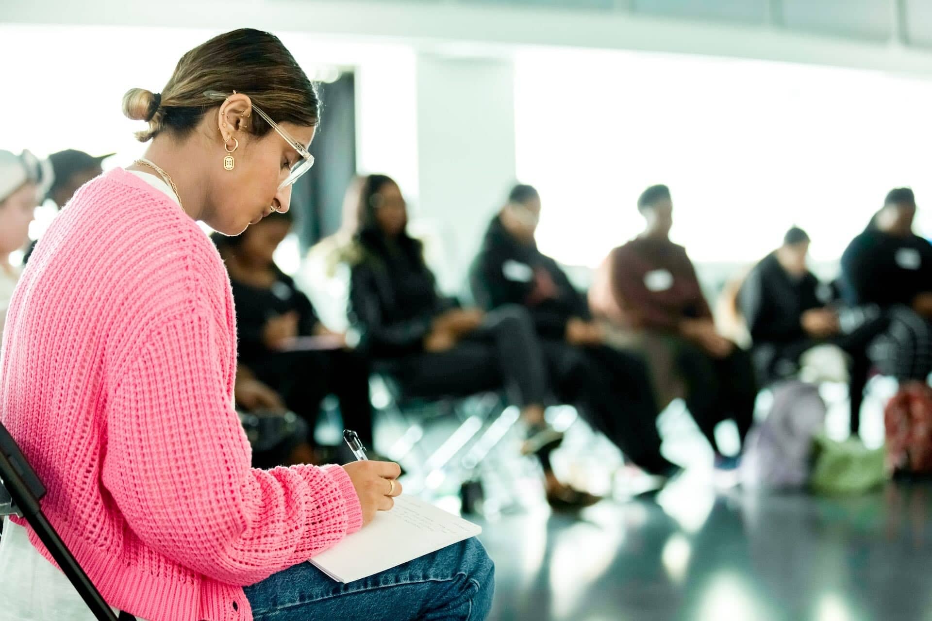 The image shows a group of people seated in a circle in what appears to be a meeting or workshop setting. The focus is on a person in the foreground wearing a bright pink sweater and glasses, writing in a notebook. The background is blurred, showing