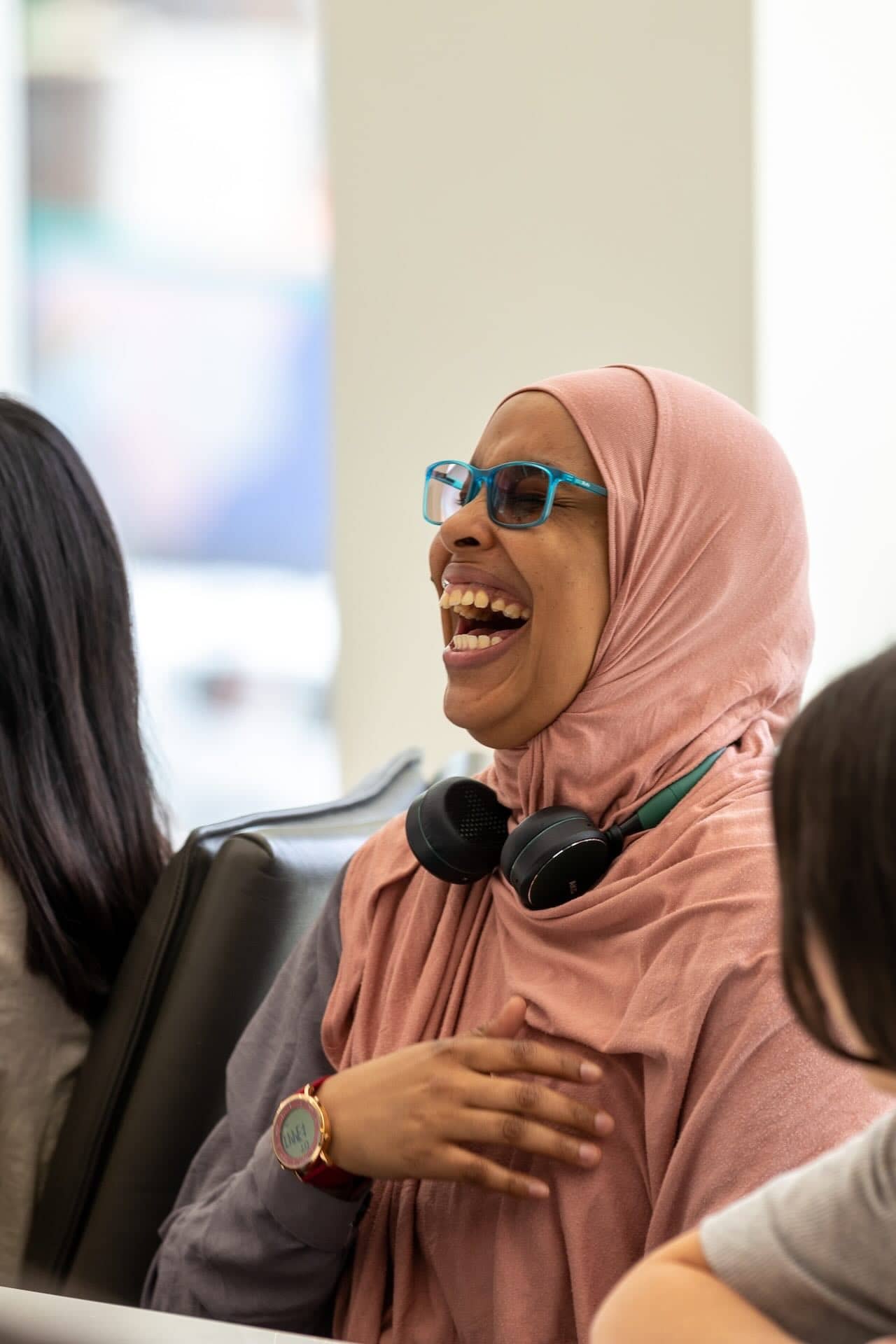The image shows a person wearing a pink hijab and glasses, laughing. They have a pair of headphones around their neck and are next to another person. The setting appears to be indoors, possibly in a casual or office environment.