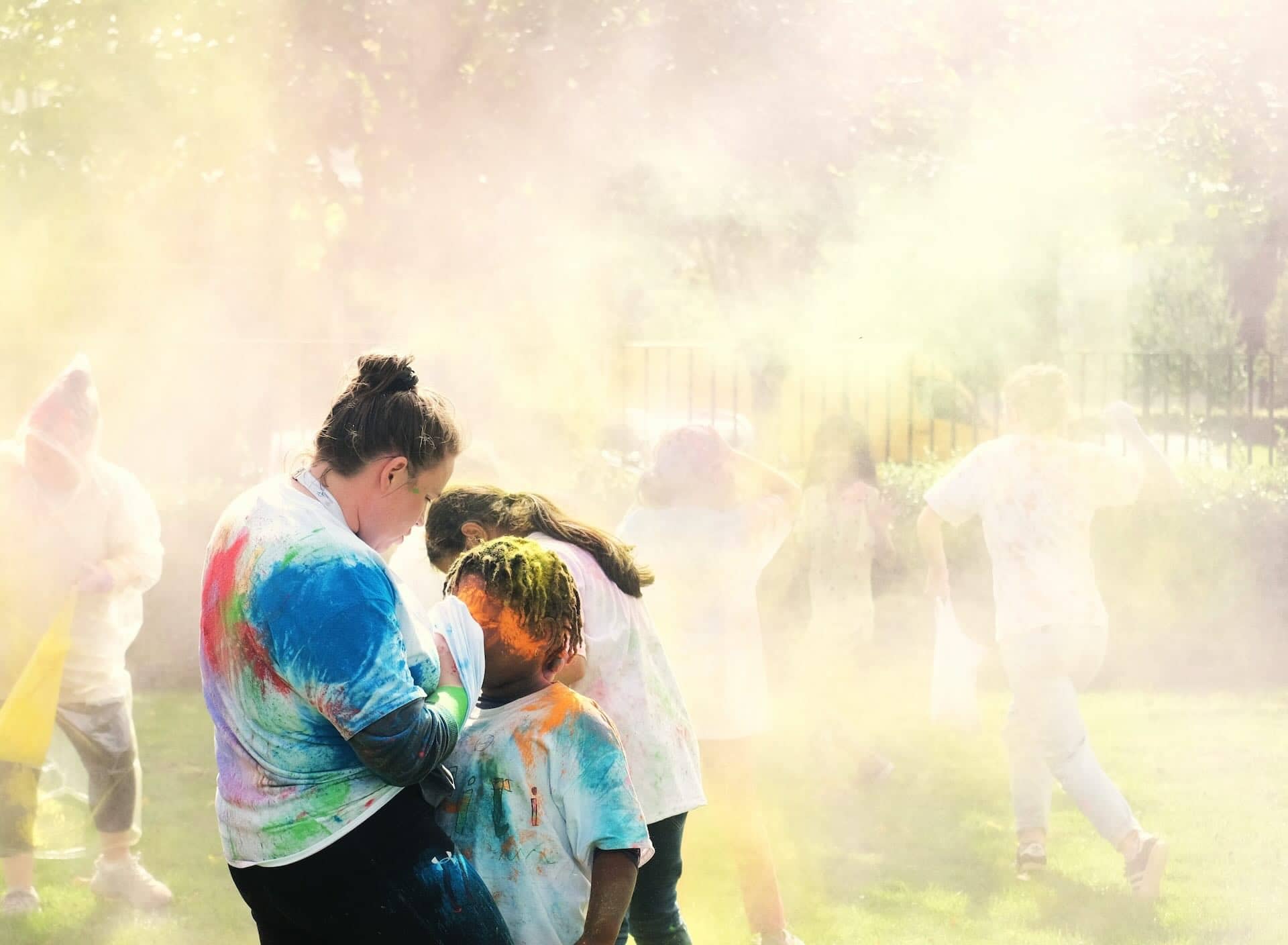 The image shows a group of people participating in what appears to be a color run or similar event. They are outdoors, with sunlight filtering through trees, and colorful powders are being thrown in the air, covering their clothes and the surroundings. Participants are wearing