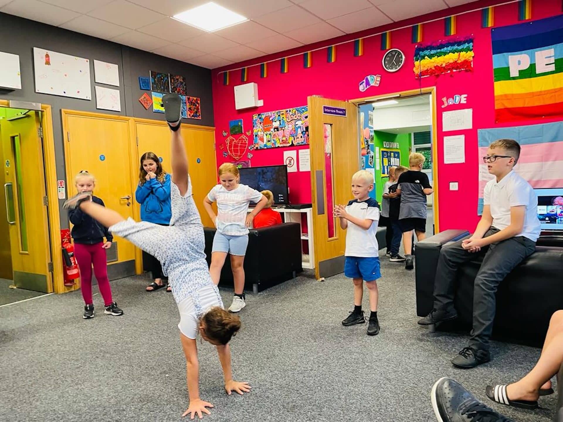 The image shows a lively scene in a colorful room, where several children are present. One child is performing a cartwheel in the center of the room. Other children are standing or sitting around, watching and engaging with each other. The walls are adorned