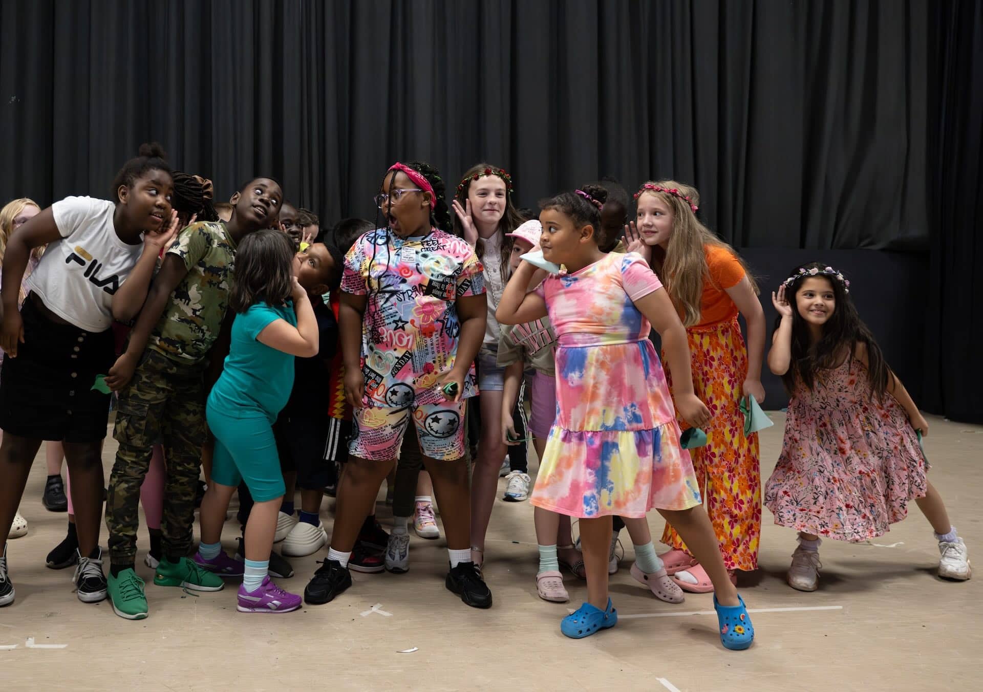 The image shows a group of children standing together in a playful pose. They are in a room with a dark curtain in the background. Some children are leaning in or cupping their ears, as if listening to a secret or sound. They are wearing