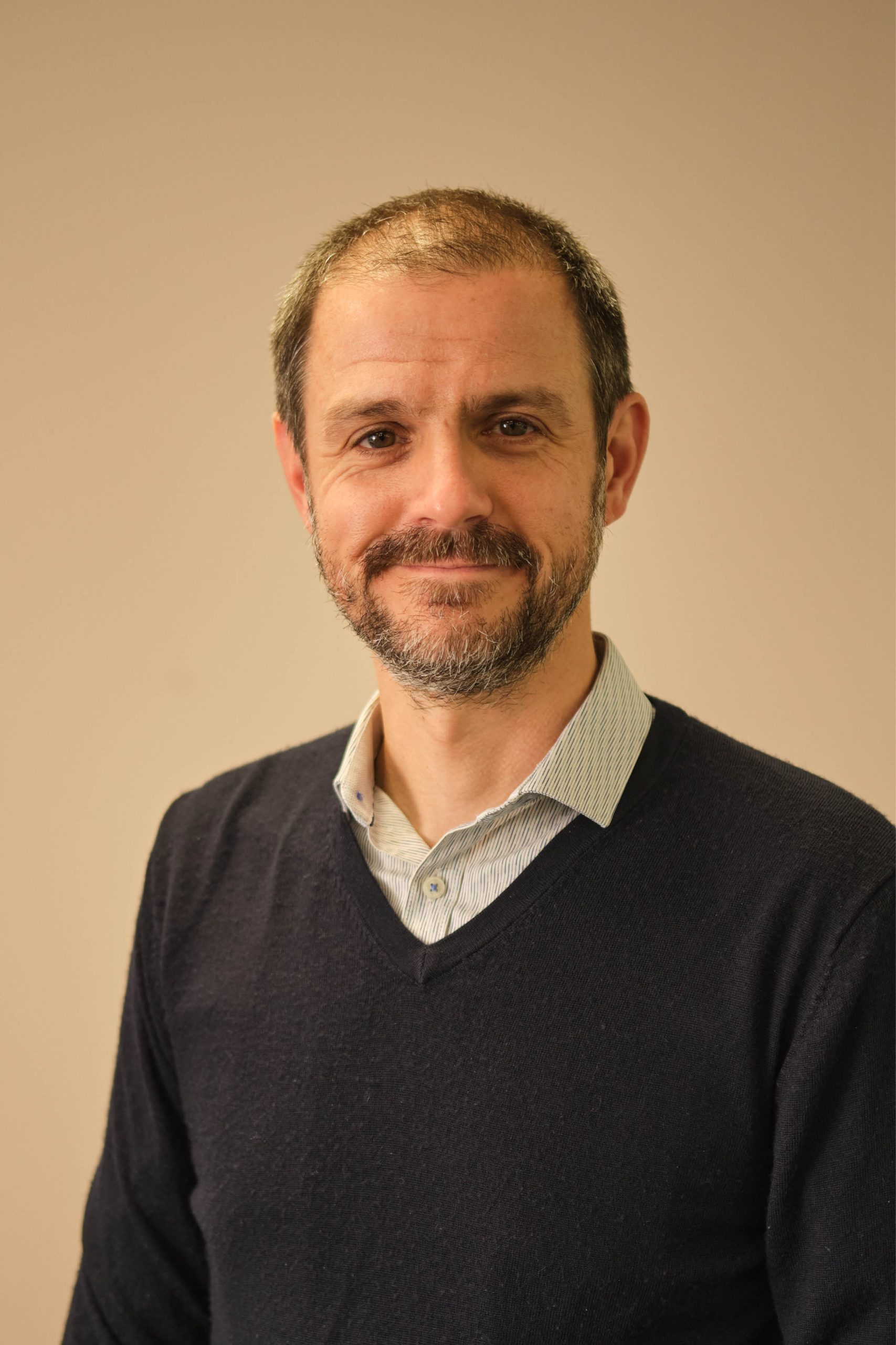 The image shows a man with a beard and short hair, wearing a dark sweater over a collared shirt, standing against a plain background.