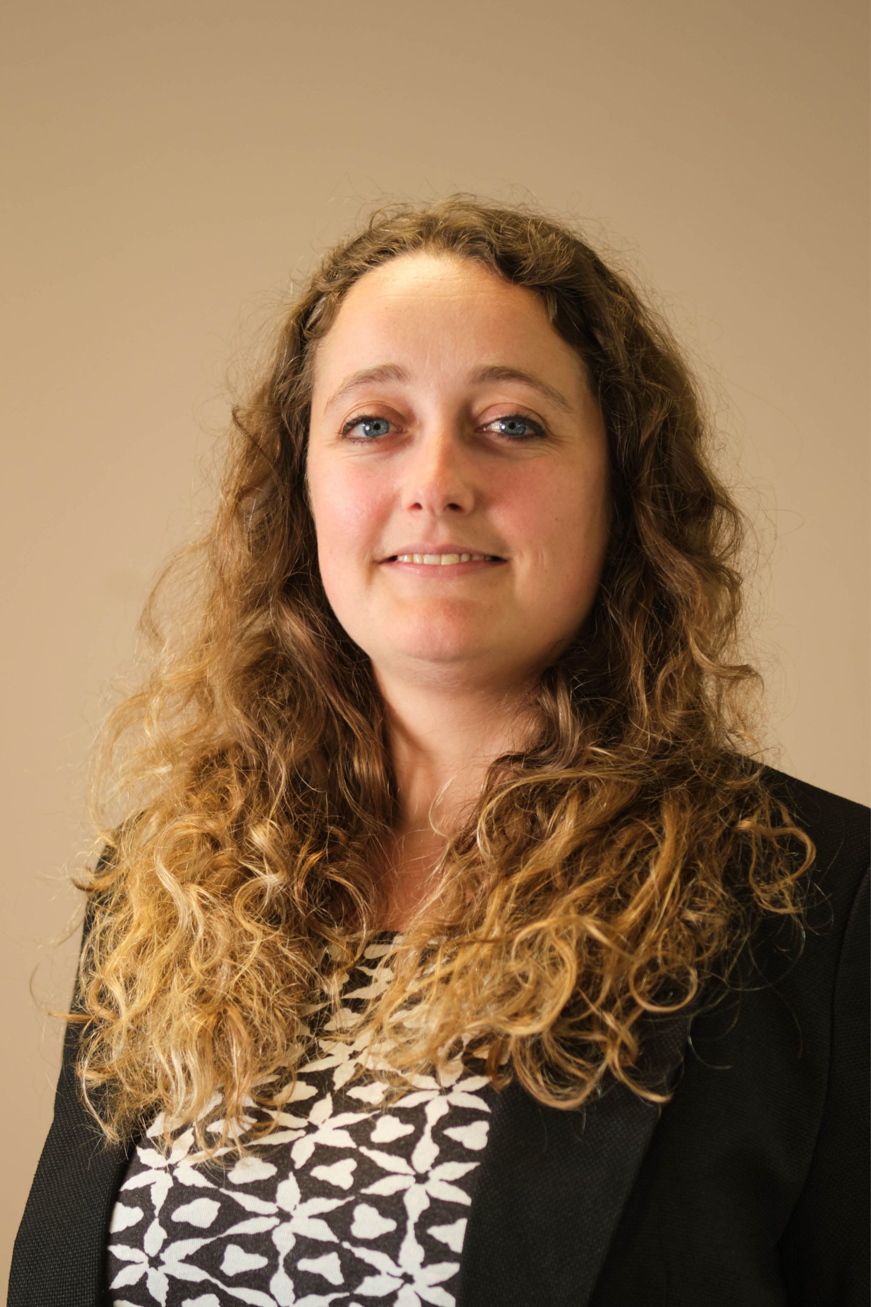 The image shows a person with long curly hair, wearing a patterned top and a black blazer. The background is plain and neutral.