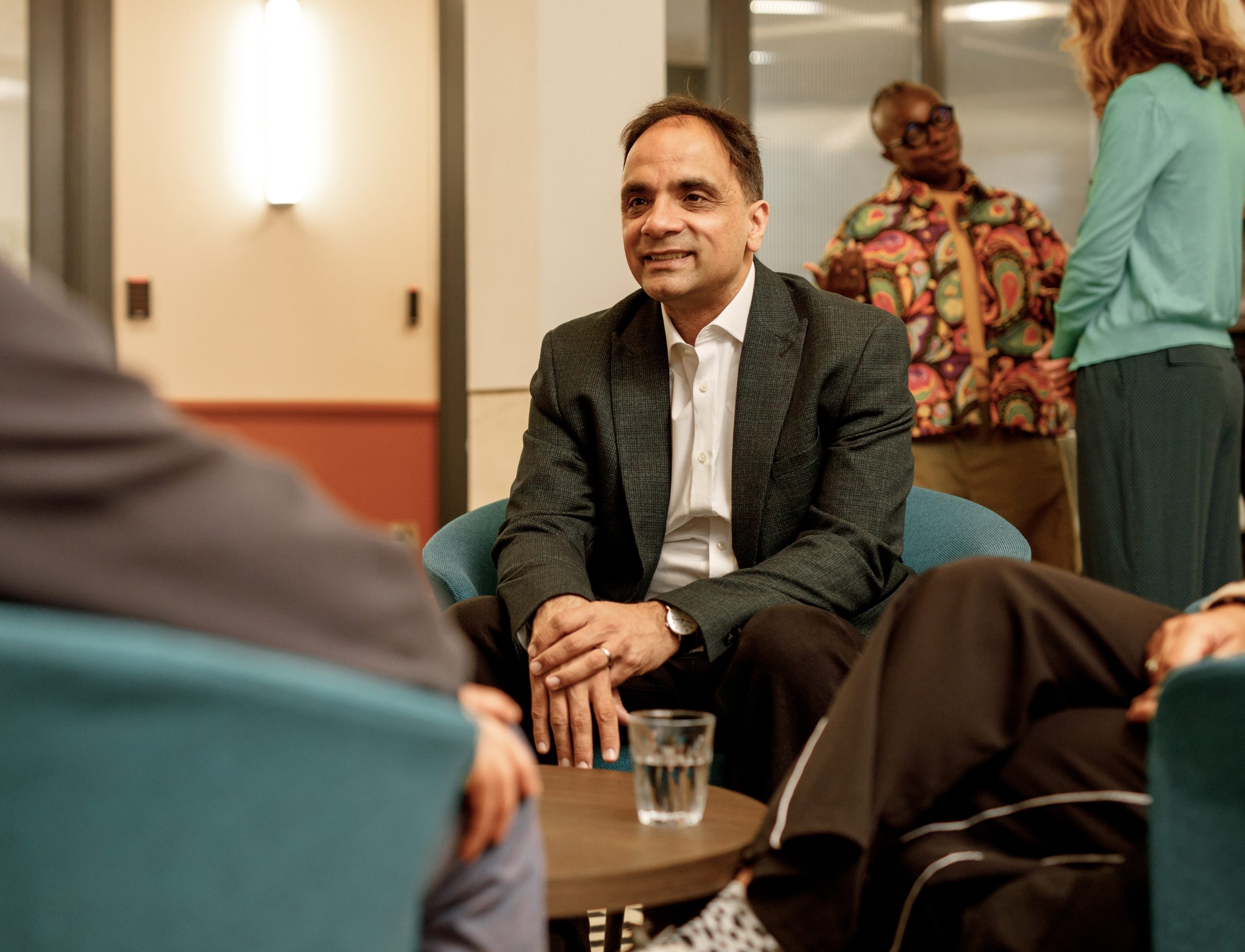 The image shows a group of people in a professional setting. A man in a suit is sitting and smiling, engaged in conversation with others. There's a glass of water on the table in front of him. In the background, two other people are standing.