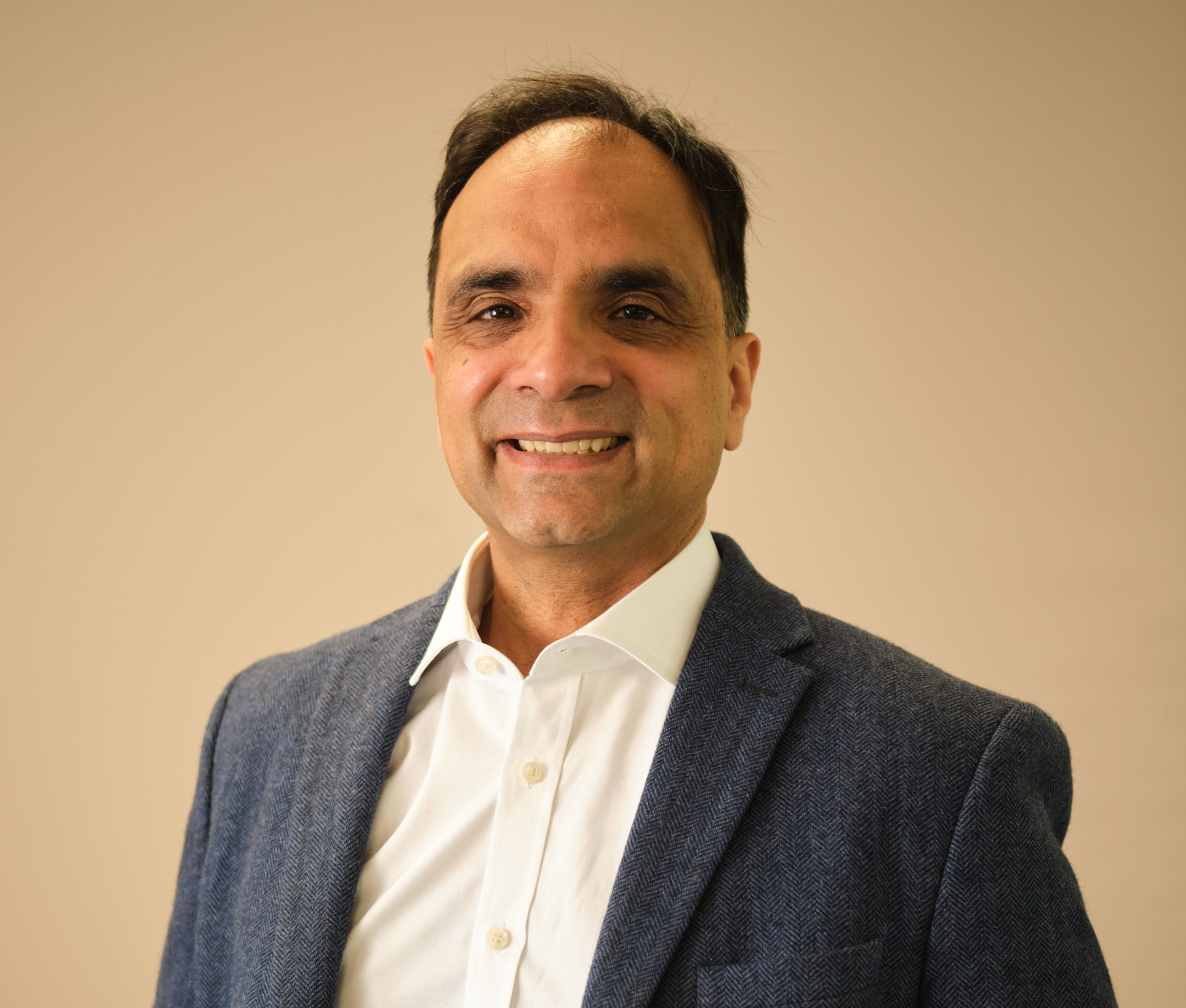 The image shows a person wearing a blazer and a white shirt, smiling against a plain background.