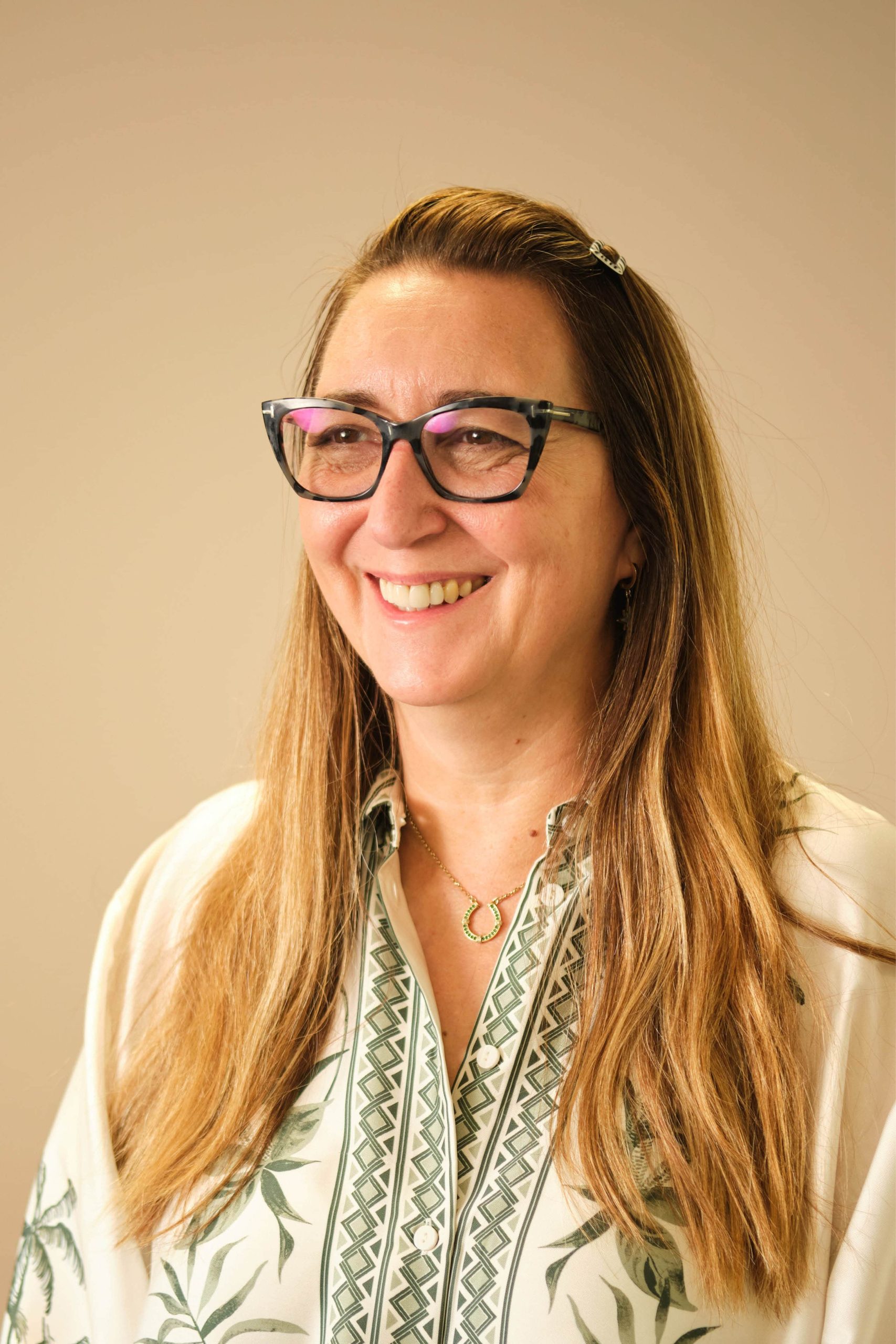 The image shows a person with long hair wearing glasses and a patterned shirt. They are smiling and looking in the direction of the light source. The background is plain and light-colored.