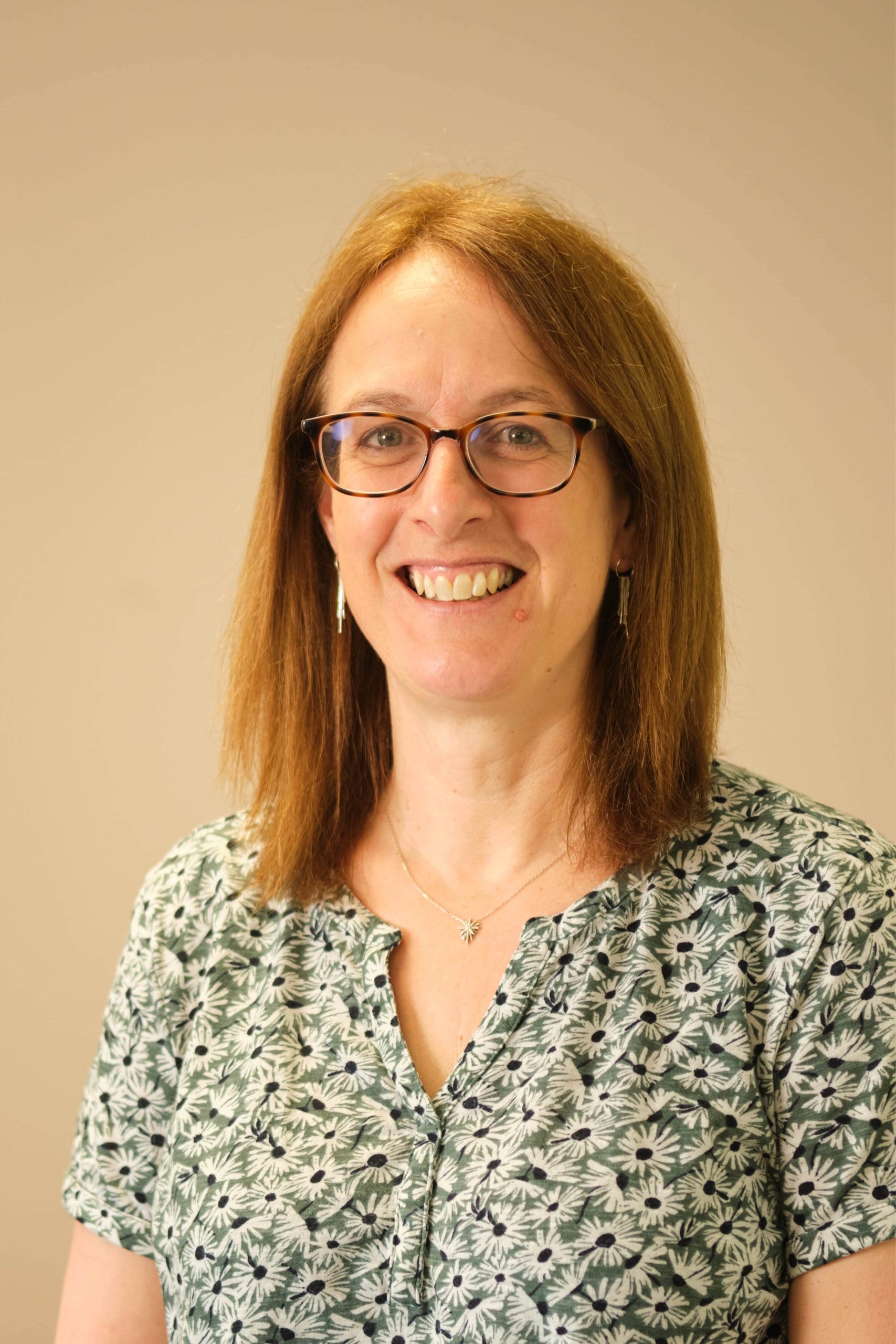 This image shows a person wearing glasses and smiling. They have shoulder-length brown hair and are wearing a patterned shirt with floral designs. There's a simple necklace with a small pendant. The background is a plain, neutral color.