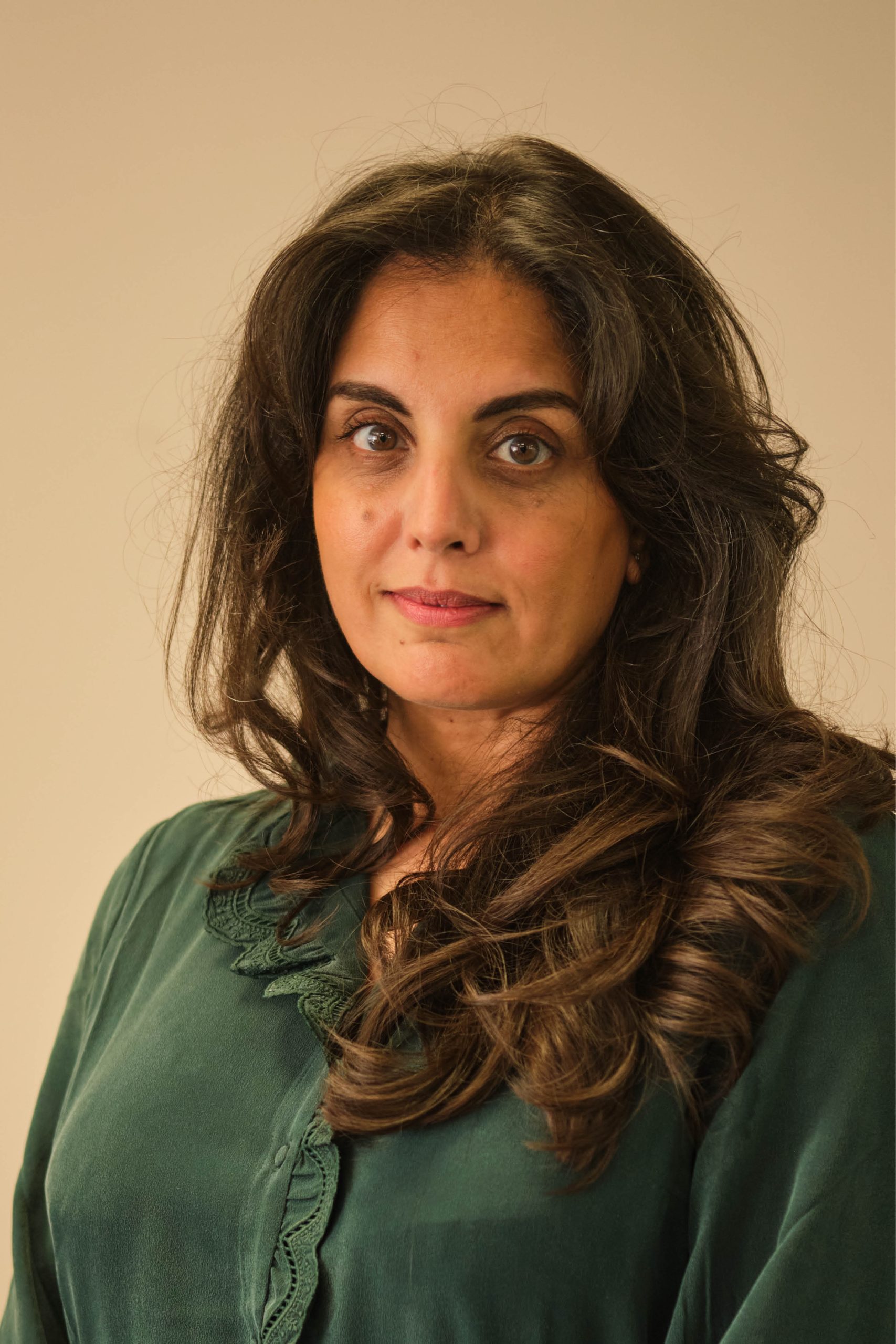 The image is a portrait of a person with long, dark hair and a serious expression. They are wearing a dark green shirt and are looking directly at the camera against a neutral background.