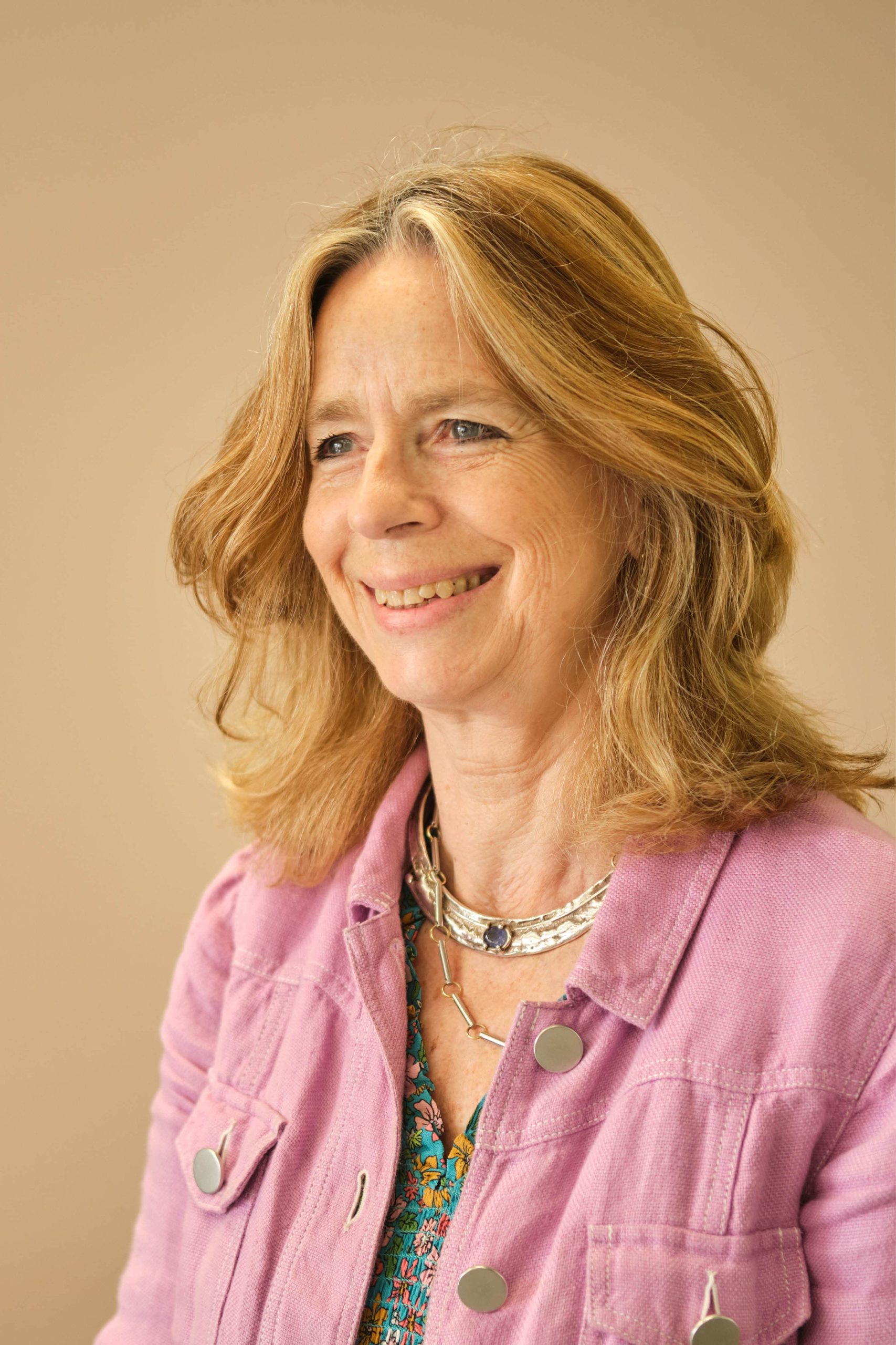 This image shows a woman with shoulder-length blonde hair, wearing a purple jacket over a patterned top. She has a necklace and is smiling. The background is a neutral color.