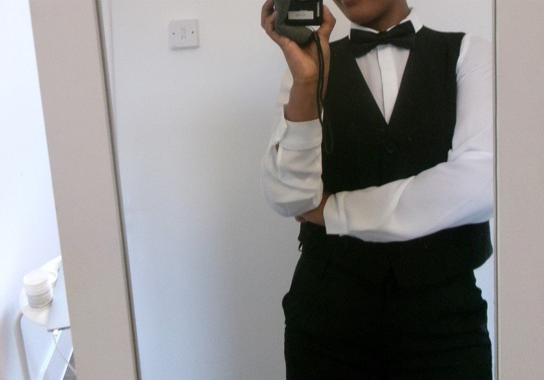 The image shows a person taking a mirror selfie. They are dressed in formal attire, wearing a white shirt, black vest, bow tie, and black pants.