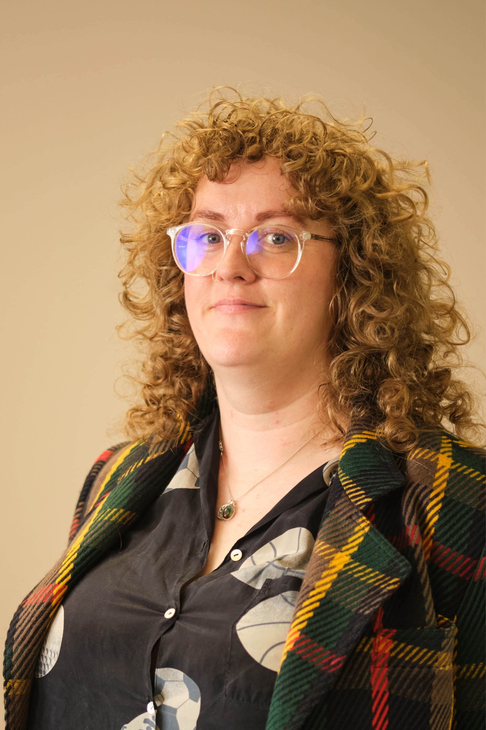 The image shows a person with curly hair wearing glasses. They are dressed in a plaid jacket and a dark shirt with a pattern, and they have a necklace around their neck. The background is a plain, neutral color.