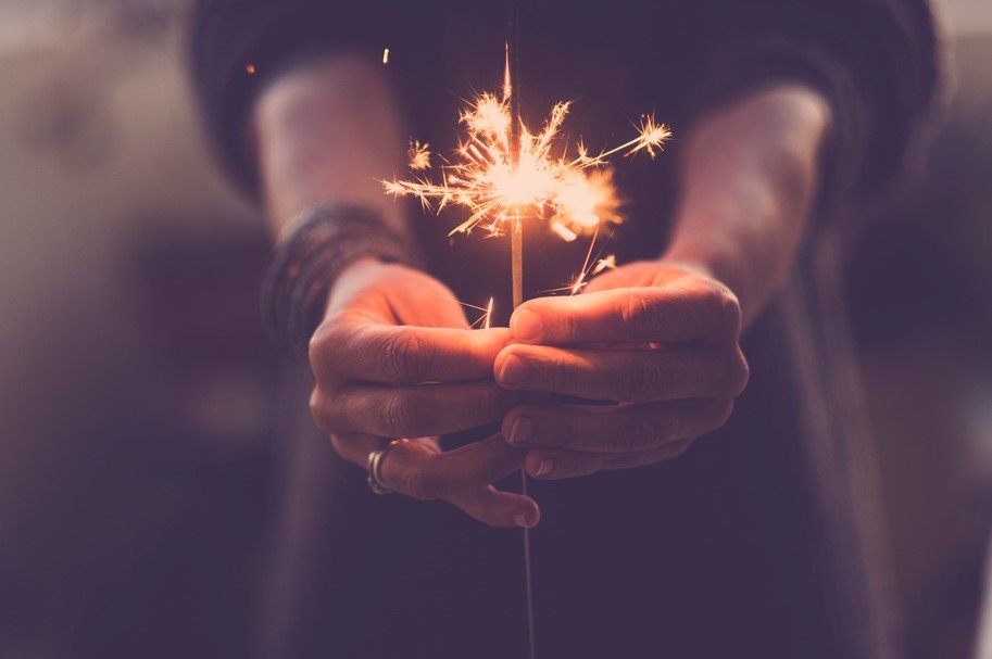 The image shows a person holding a lit sparkler. The focus is on the hands and the sparkling effect of the sparkler. The background is blurred, giving a warm and festive atmosphere.