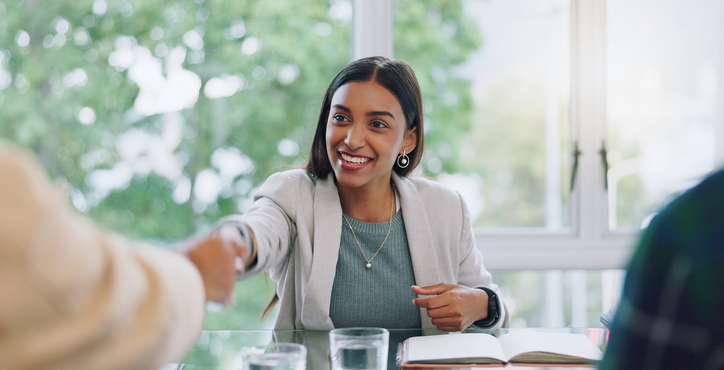 The image shows a woman sitting at a table, smiling and shaking hands with someone. She is wearing a light-colored blazer and a blue top, and there are glass cups and an open notebook on the table. The setting appears to be an office or