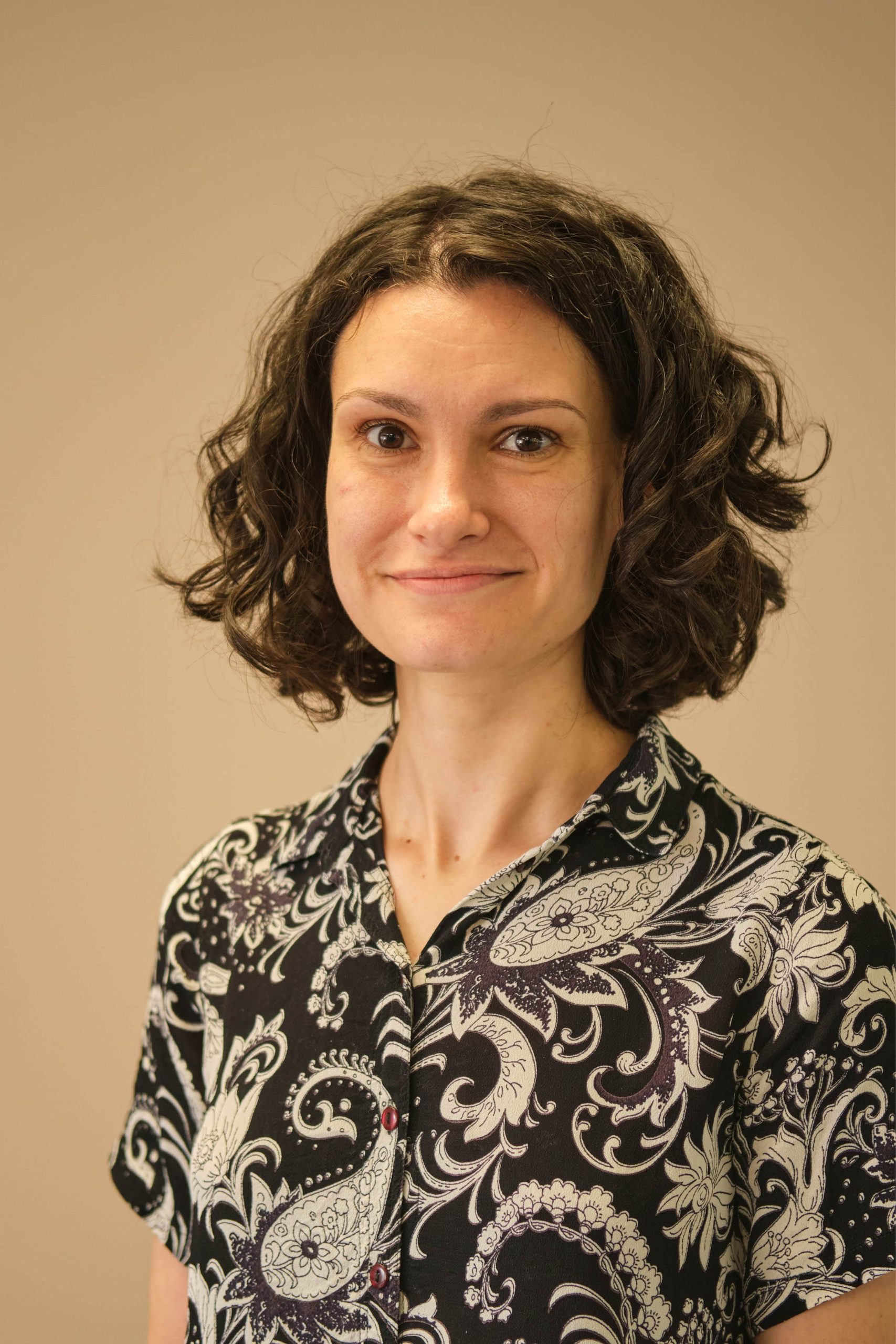 The image shows a person with short, curly hair, wearing a patterned shirt featuring floral and paisley designs. The background is a plain, neutral color.