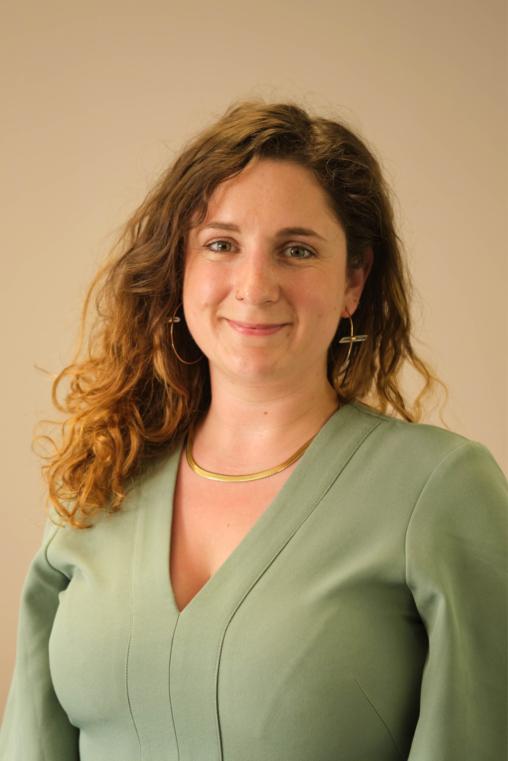 The image shows a person with long, wavy hair, wearing a green top and gold jewelry, including hoop earrings and a necklace. They are smiling against a plain background.