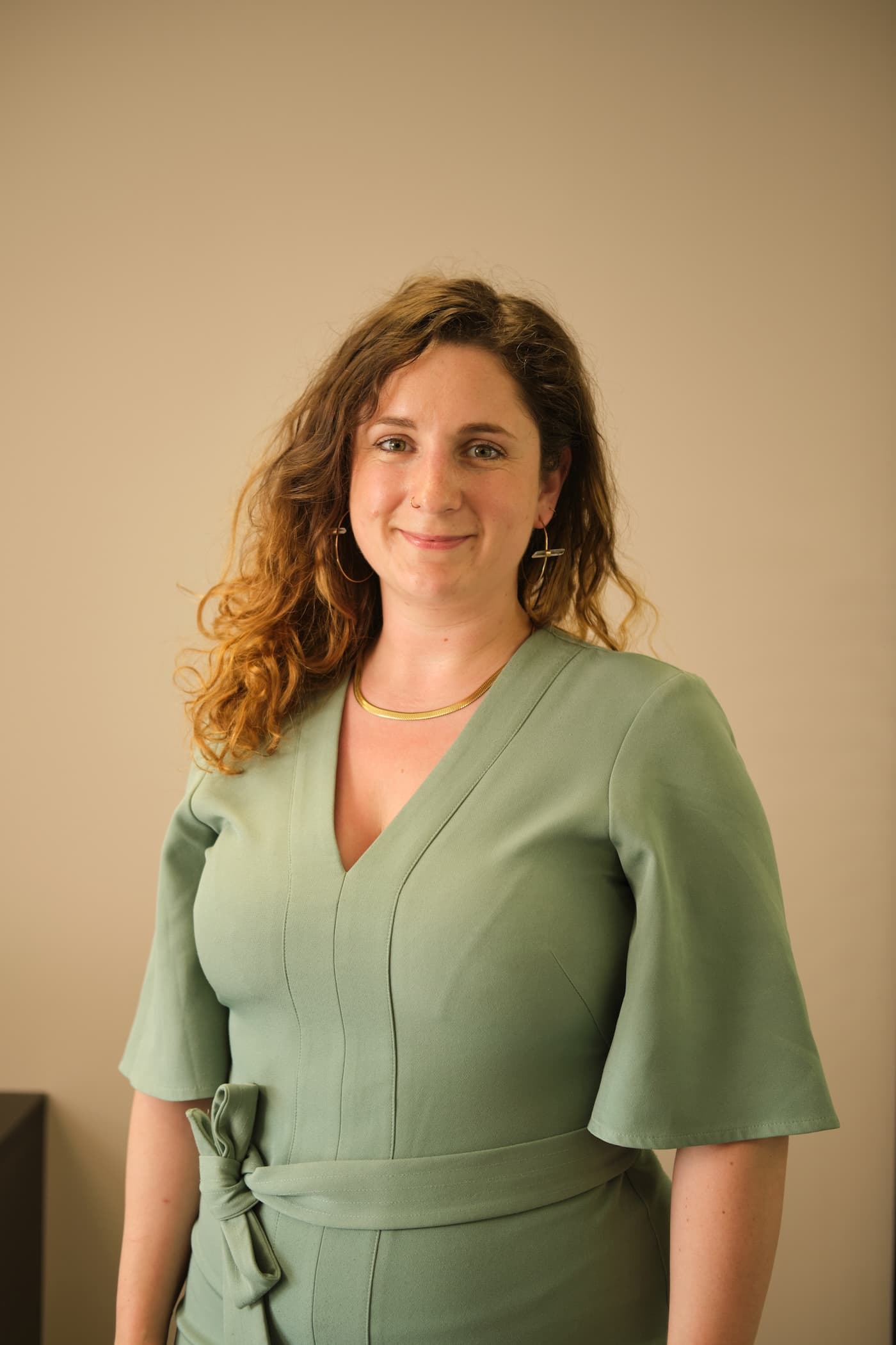 This image shows a person with long, wavy hair wearing a green dress. The dress has a tie at the waist and elbow-length sleeves. The person is smiling and wearing earrings and a necklace.