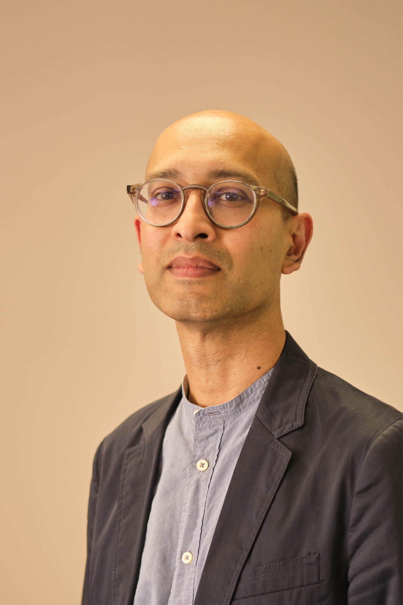 The image shows a person wearing glasses, a blue shirt with buttons, and a dark blazer. The background is a plain, neutral color.