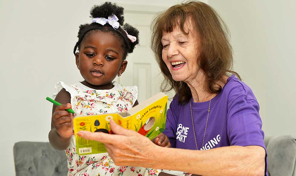 The image shows a young child and an older woman who appear to be enjoying a moment together. The woman is holding a colorful children's book, and the child is holding a green crayon, seemingly engaged in a reading or drawing activity. Both appear to