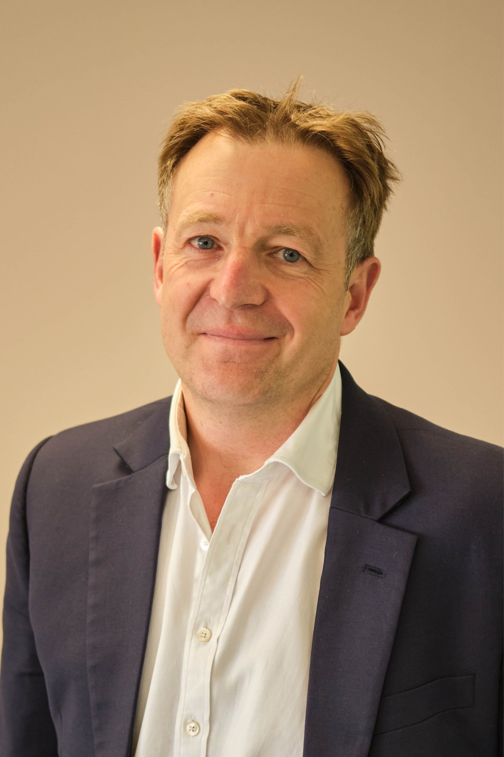 The image shows a man wearing a dark suit jacket over a white dress shirt. He's posing against a plain background and is smiling slightly.