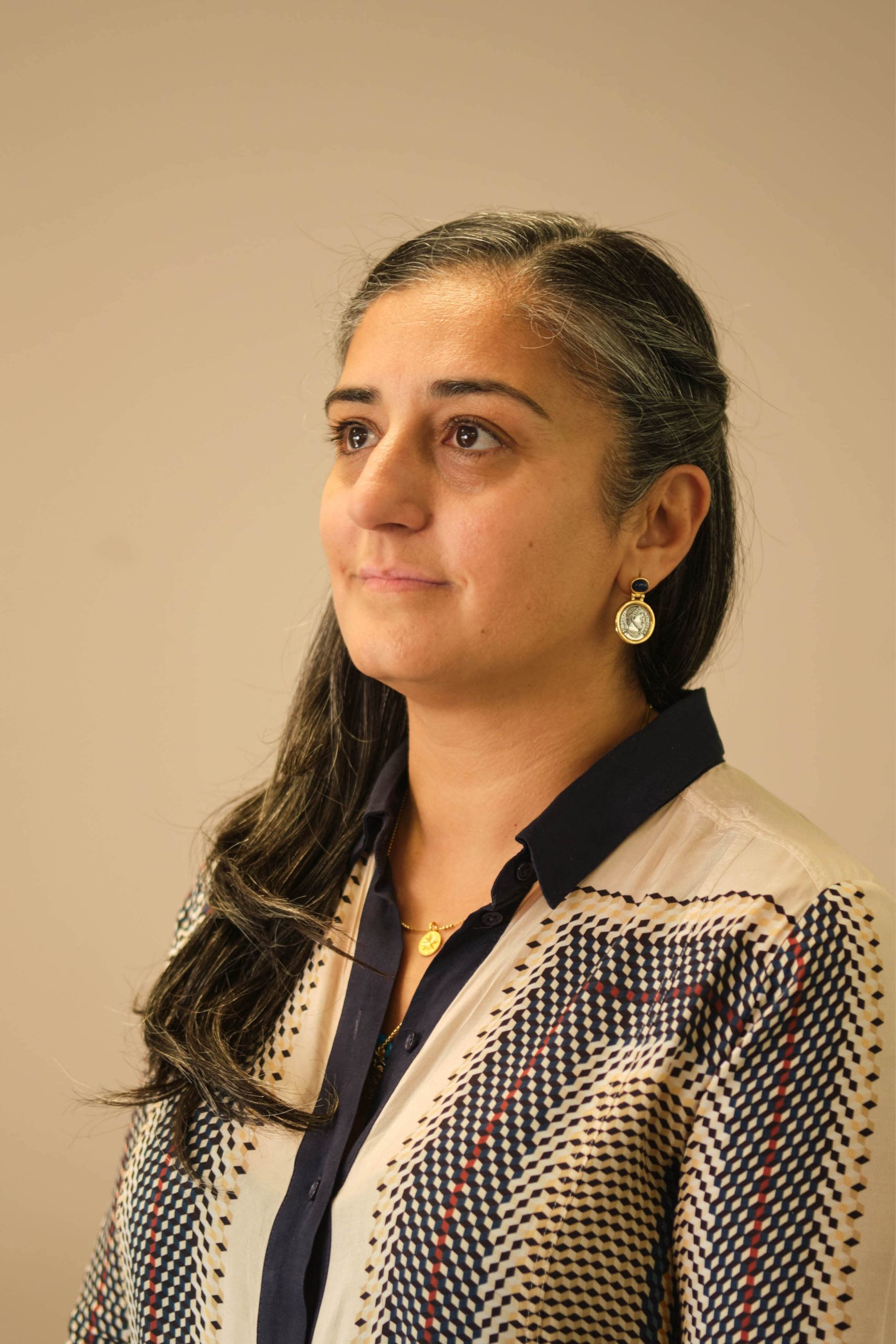 The image shows a person with long, dark hair, wearing a patterned blouse and gold earrings. The background is a plain, neutral color.