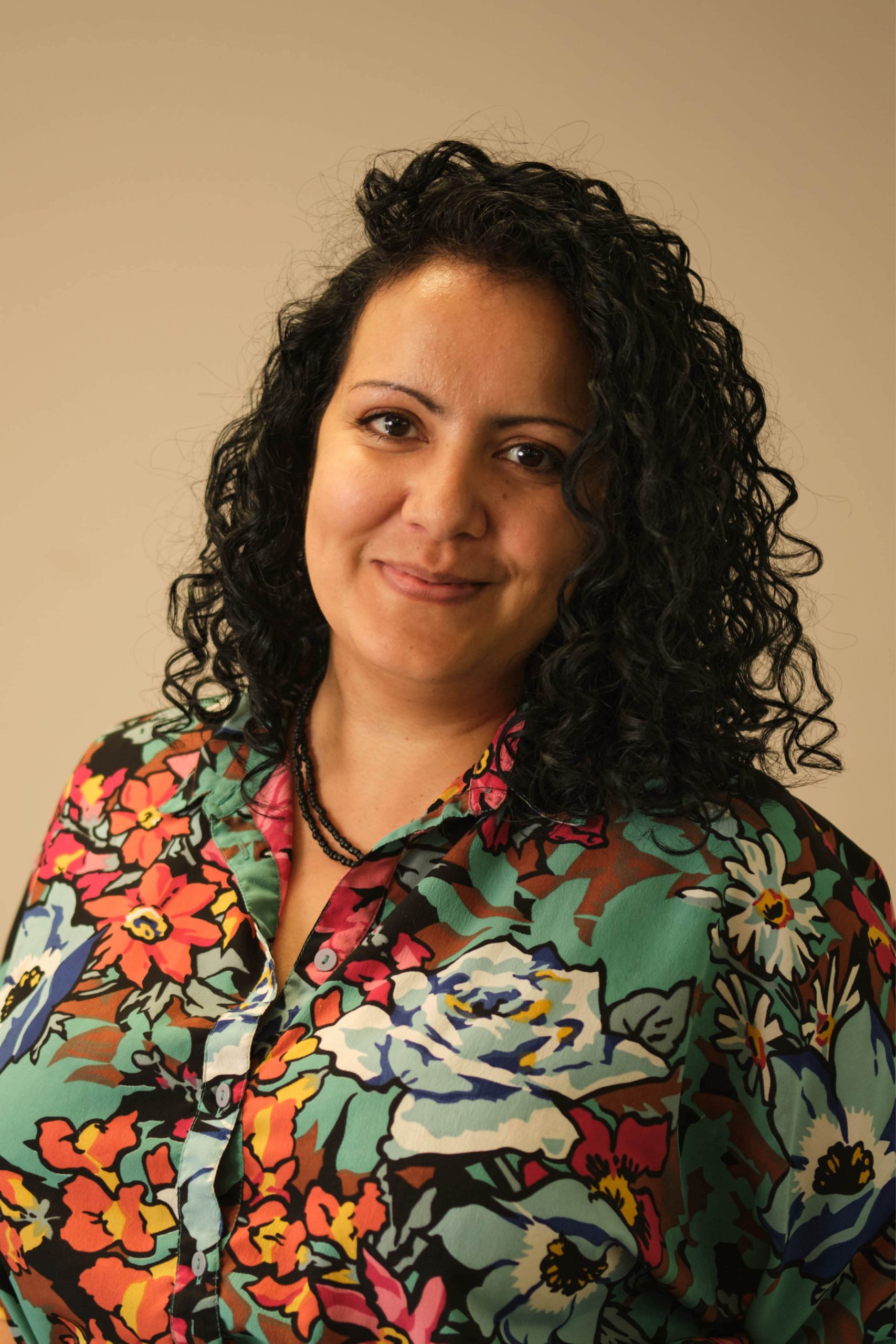 The image shows a person with curly hair wearing a colorful, floral-patterned shirt. The background is plain and the person is smiling gently, looking directly at the camera.