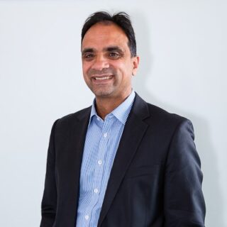 A photo of Anand Shukla, Chief Executive, wearing a Navy suit and blue shirt, and smiling