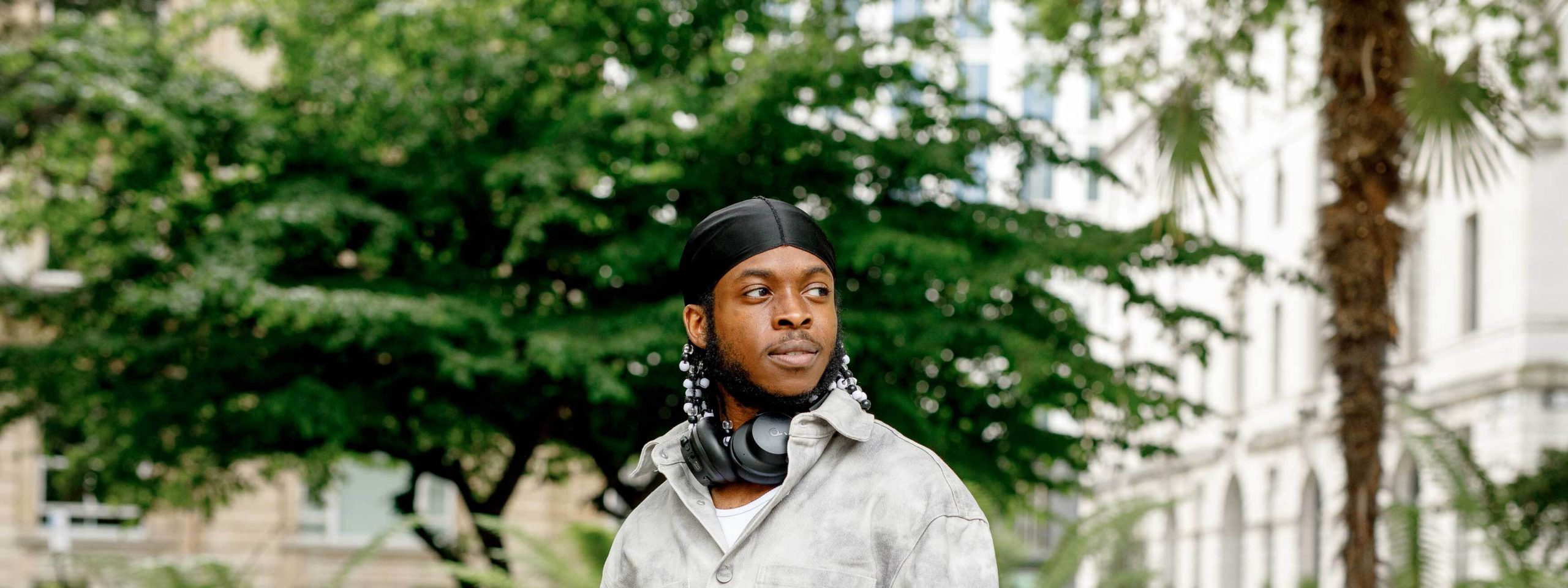 The image shows a person wearing a black headscarf and large headphones around their neck. They are dressed in a light-colored jacket and standing outdoors. In the background, there are trees and a building.
