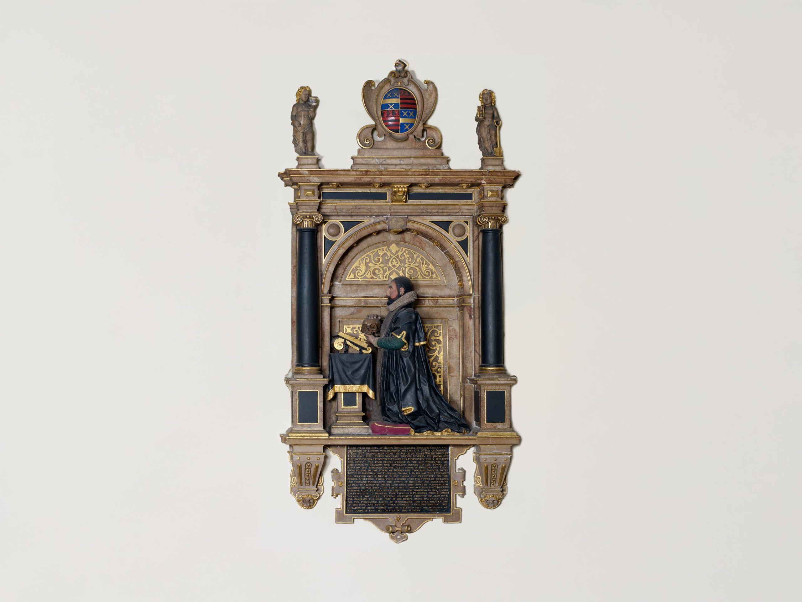 The image shows an ornate wall plaque or memorial, featuring a relief sculpture. The design includes a figure in a kneeling position, dressed in traditional robes, positioned in front of an elaborately decorated backdrop with columns and arches. The top of the plaque