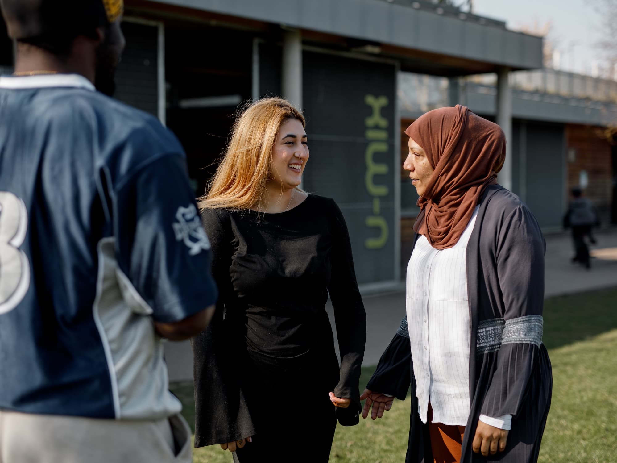 The image shows three people outdoors, interacting with each other. One person is wearing a blue sports jersey, another person is in a black outfit, and the third person is wearing a brown headscarf and a dark jacket. They appear to be talking