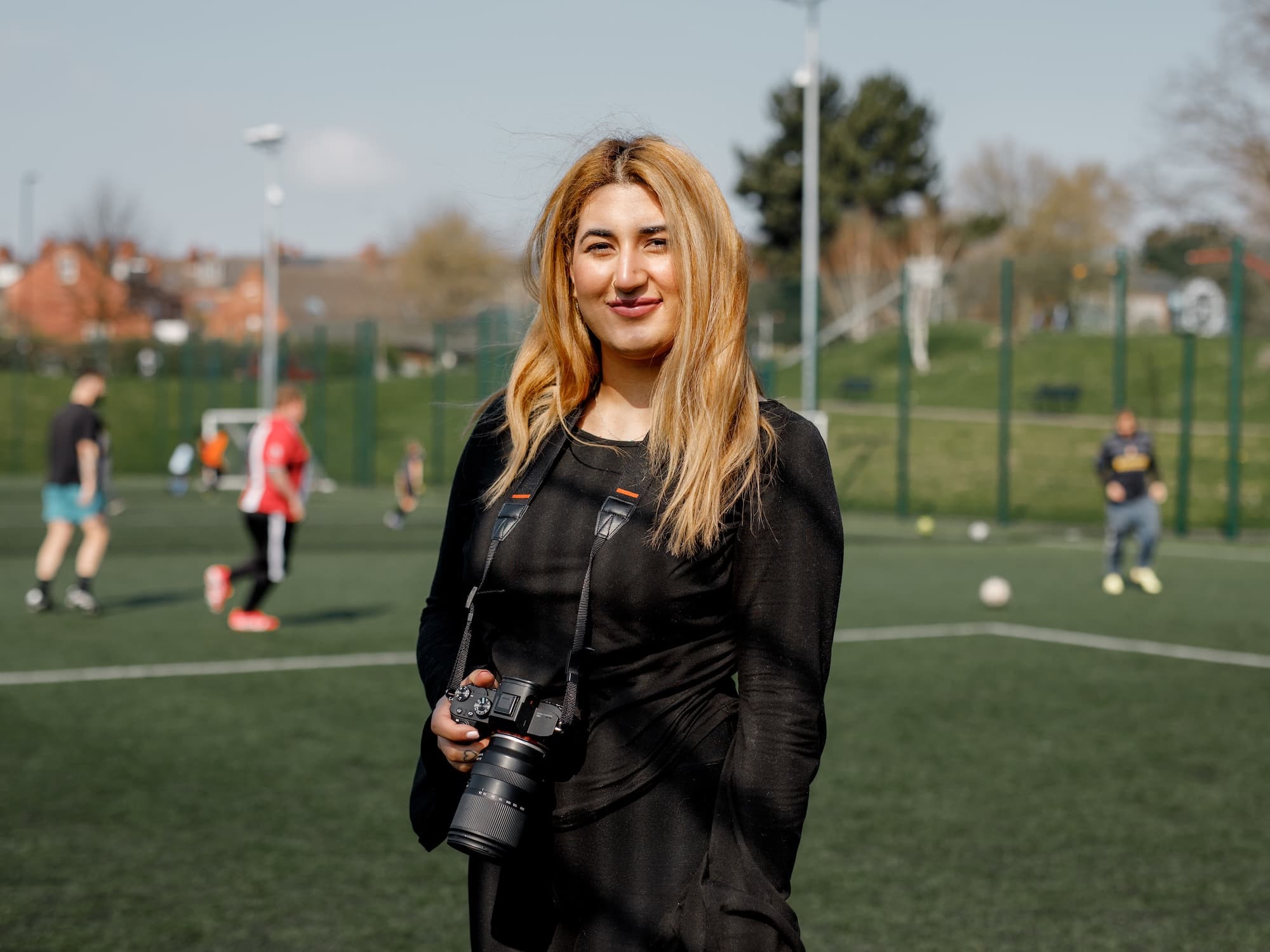 The image shows a person standing on a soccer field holding a camera. There are people playing soccer in the background. The setting appears to be outdoors, with a clear sky and some trees and buildings visible.
