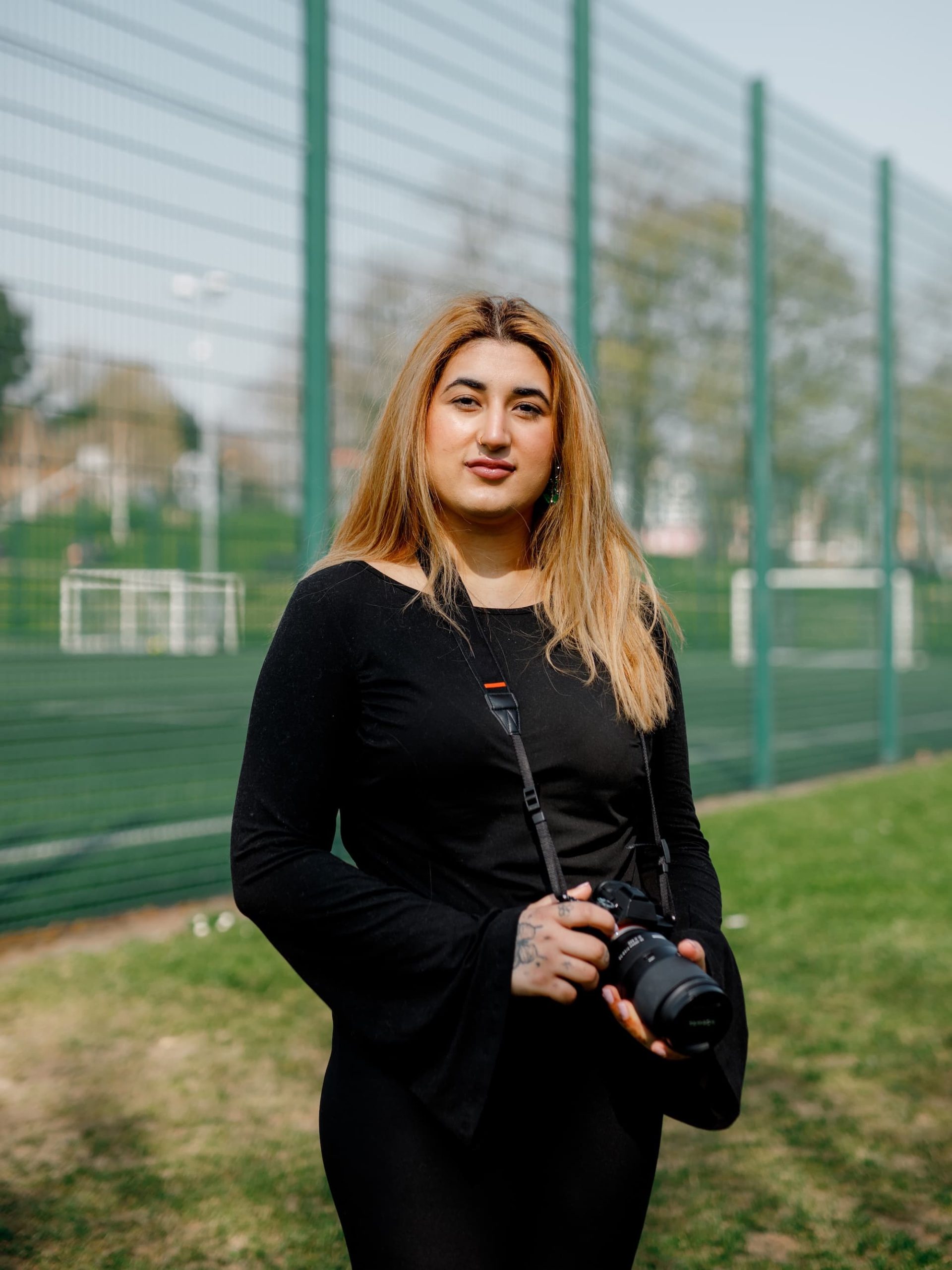 The image shows a person standing outdoors on a grassy area in front of a wire fence. They are holding a camera and wearing a black outfit. The background includes a sports field or similar area with goalposts and some trees.