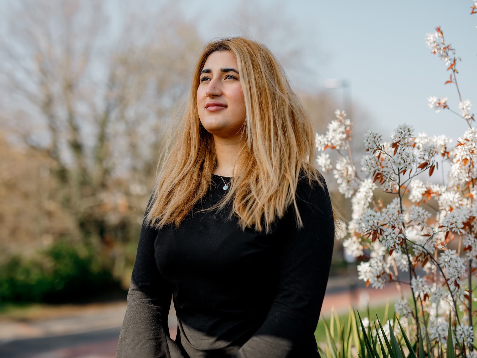 The image shows a person with long, light brown hair standing outside near some flowering plants. The person is wearing a black top and is looking upwards. The background is slightly blurred, showing trees and a clear sky.