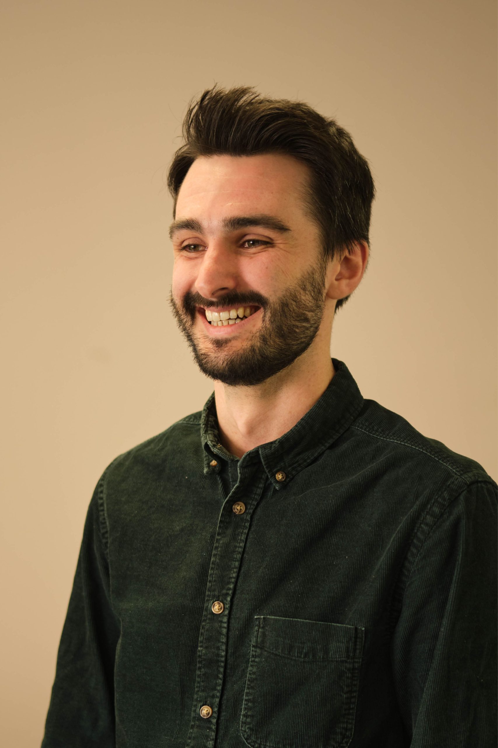 The image shows a man with dark hair and a beard, smiling and looking to his left. He is wearing a dark green corduroy shirt. The background is plain and beige.