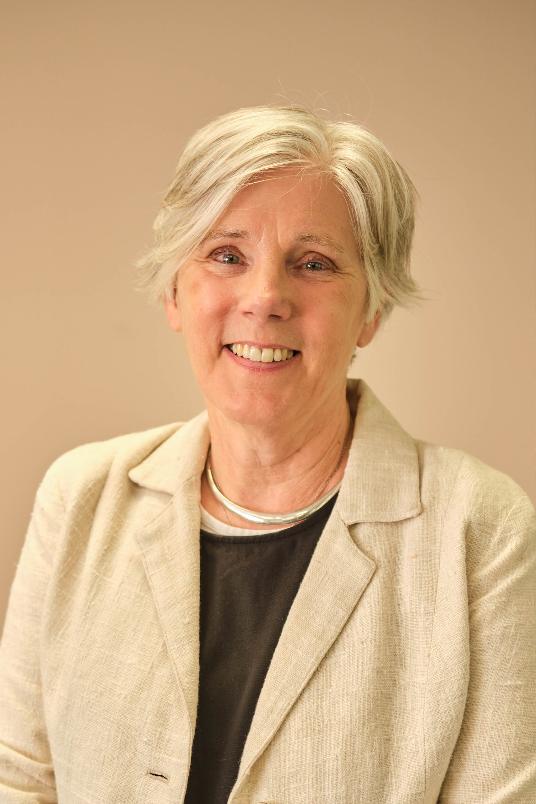 The image shows a person with short gray hair, wearing a light-colored blazer over a black top, and smiling at the camera.
