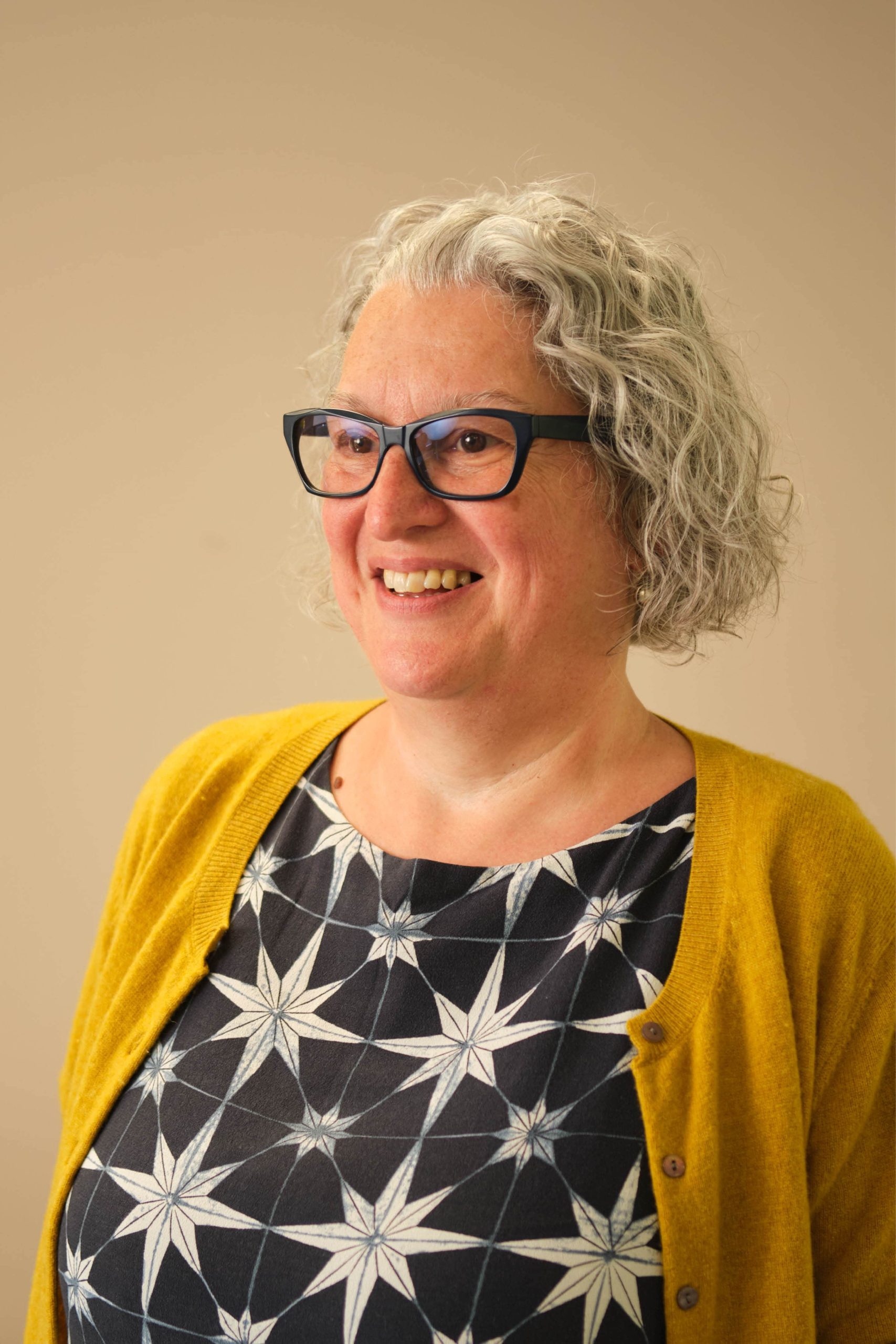 The image shows a person with gray curly hair wearing glasses, a yellow cardigan, and a patterned top with star-like designs. They are smiling and looking off to the side against a plain background.