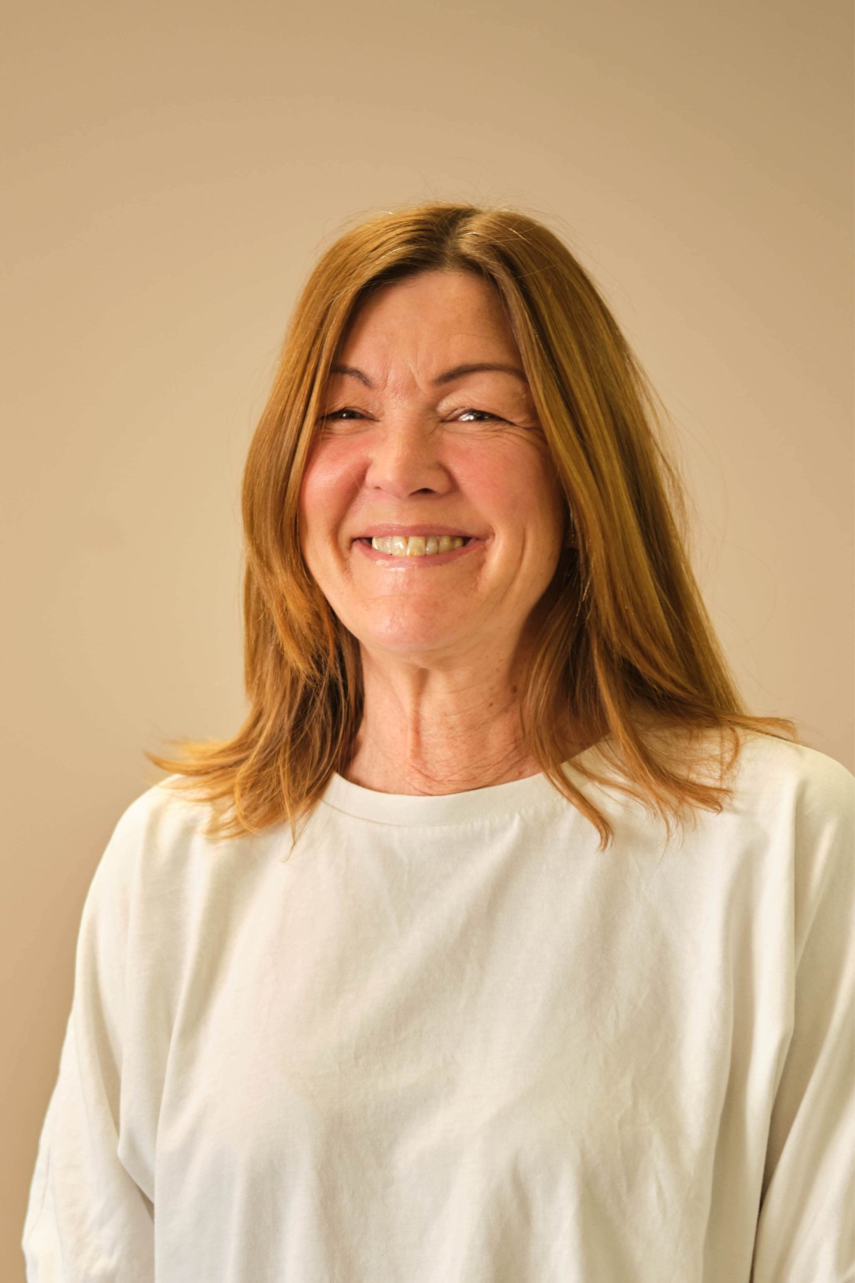 The image shows a person with shoulder-length brown hair wearing a white shirt. They are smiling and the background is a plain beige color.