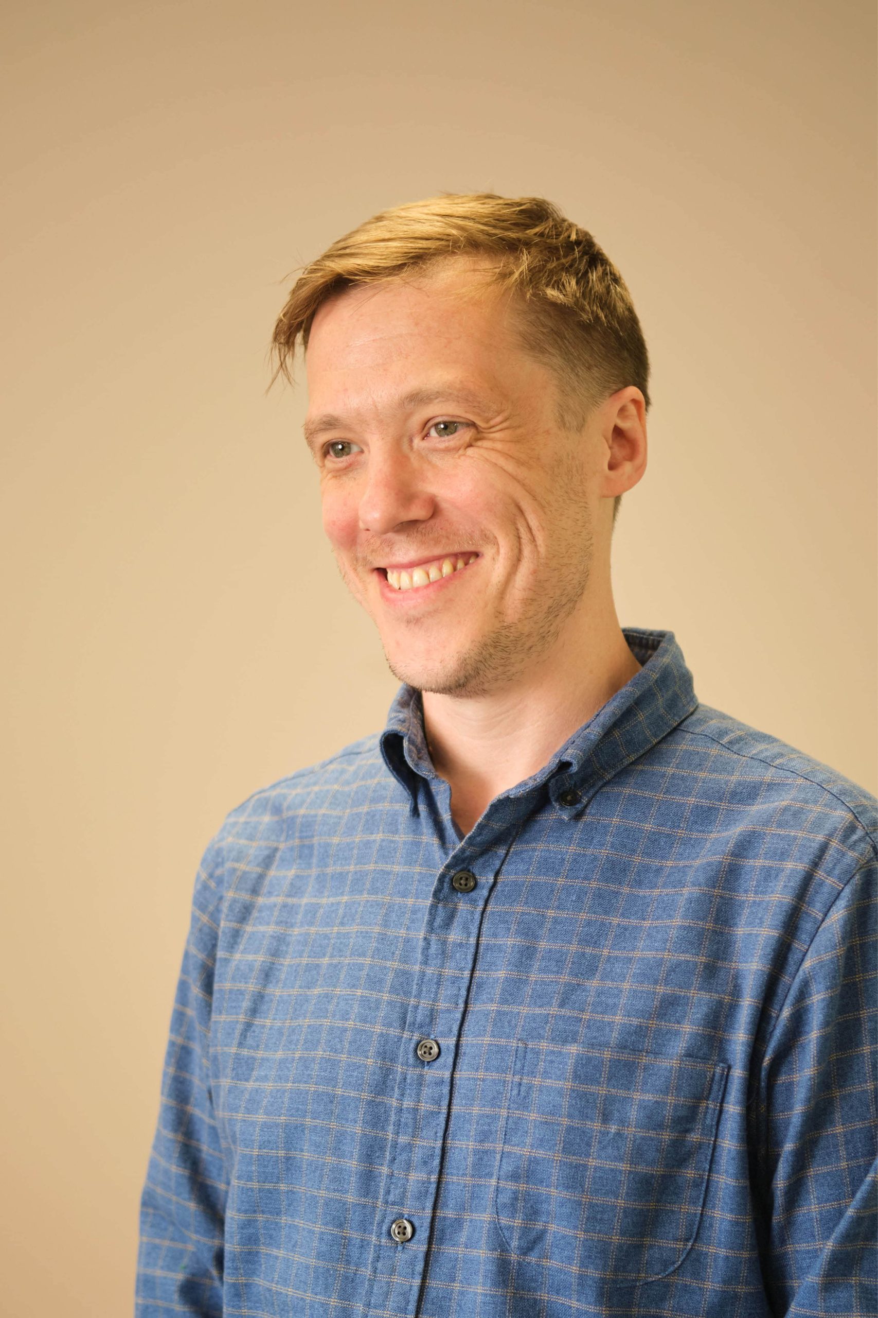 The image shows a person smiling, wearing a blue checkered shirt, against a plain background.